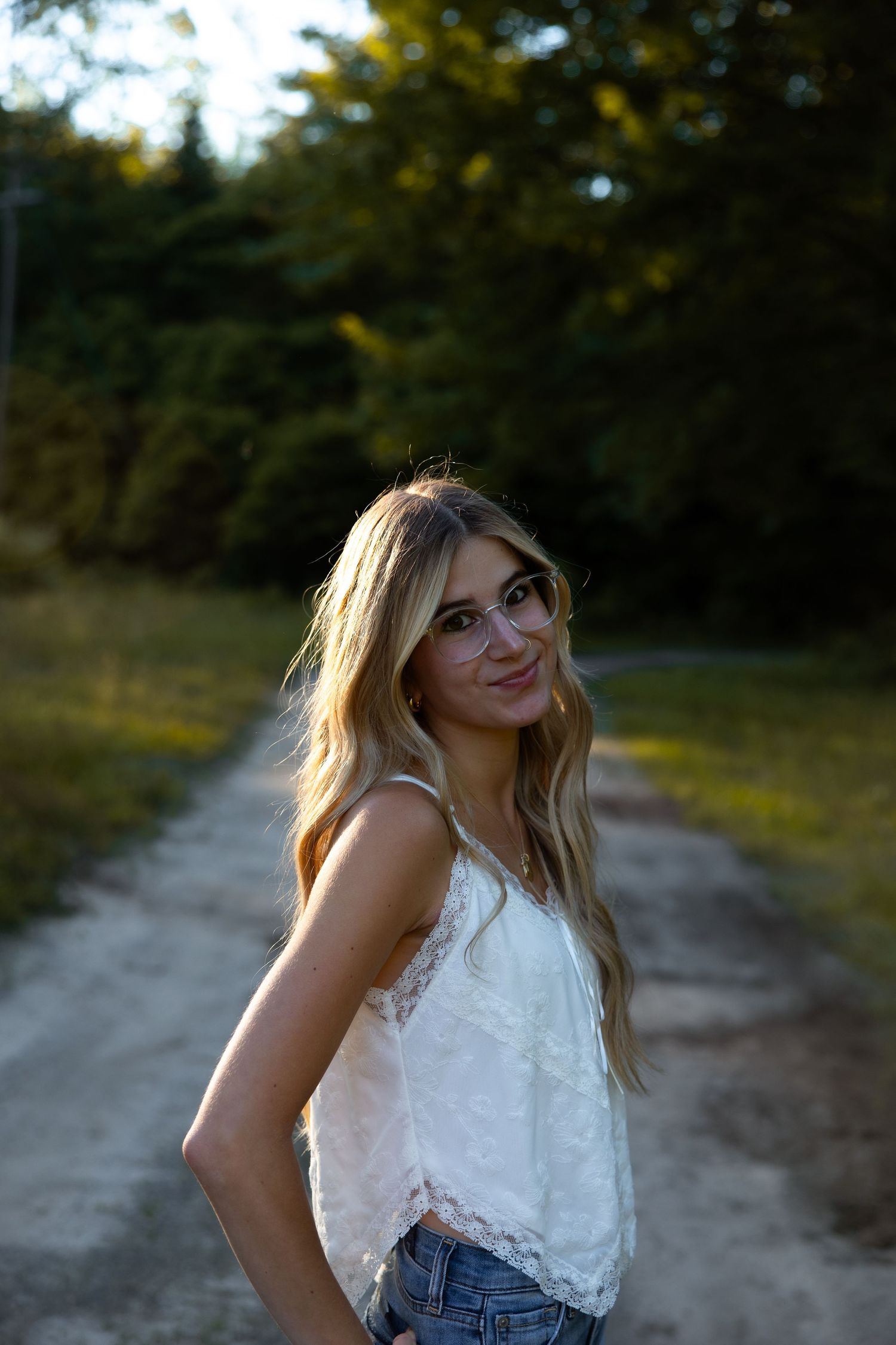 Young blonde woman with glasses in white lace top smiling over shoulder on a sunlit outdoor dirt path at dusk.