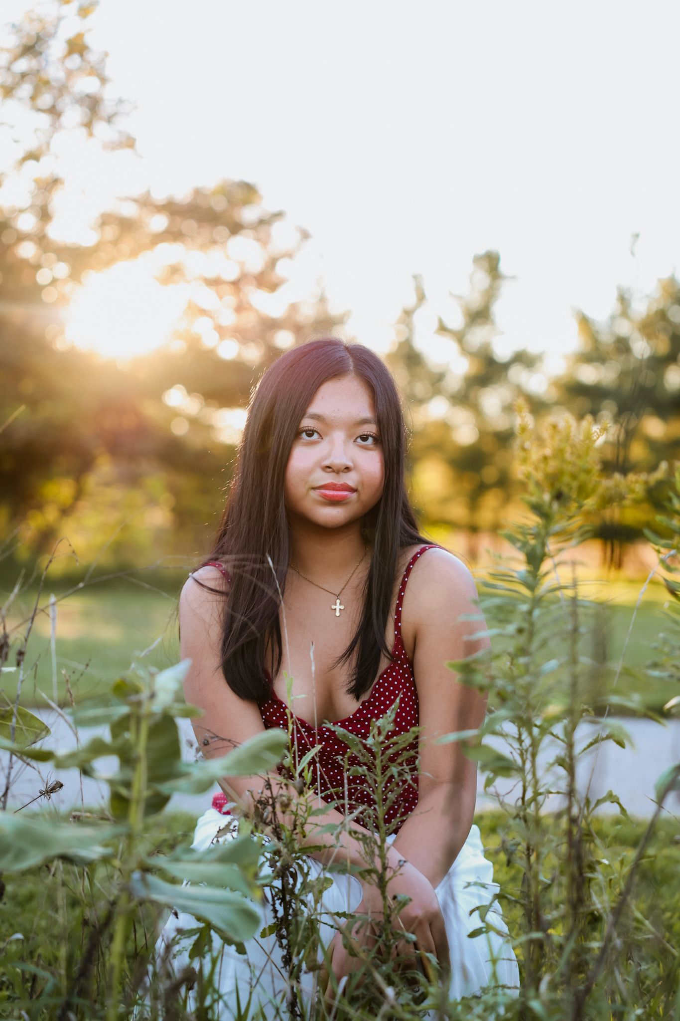 Young Asian woman in red polka dot top kneels among wildflowers in a golden sunlit field.