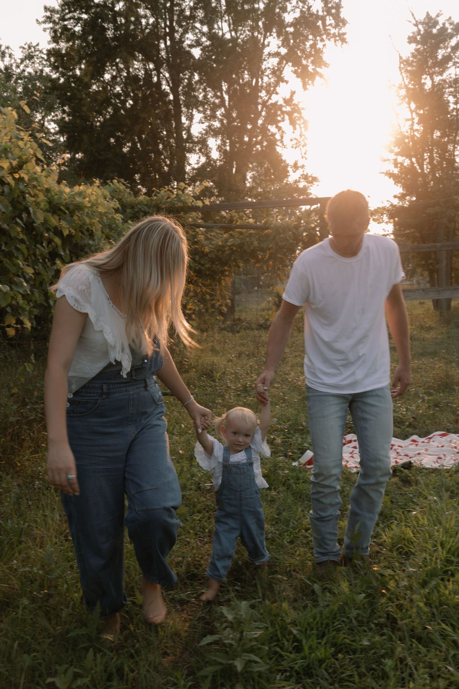 Young family walking hand-in-hand with toddler in denim overalls through lush garden at sunset.