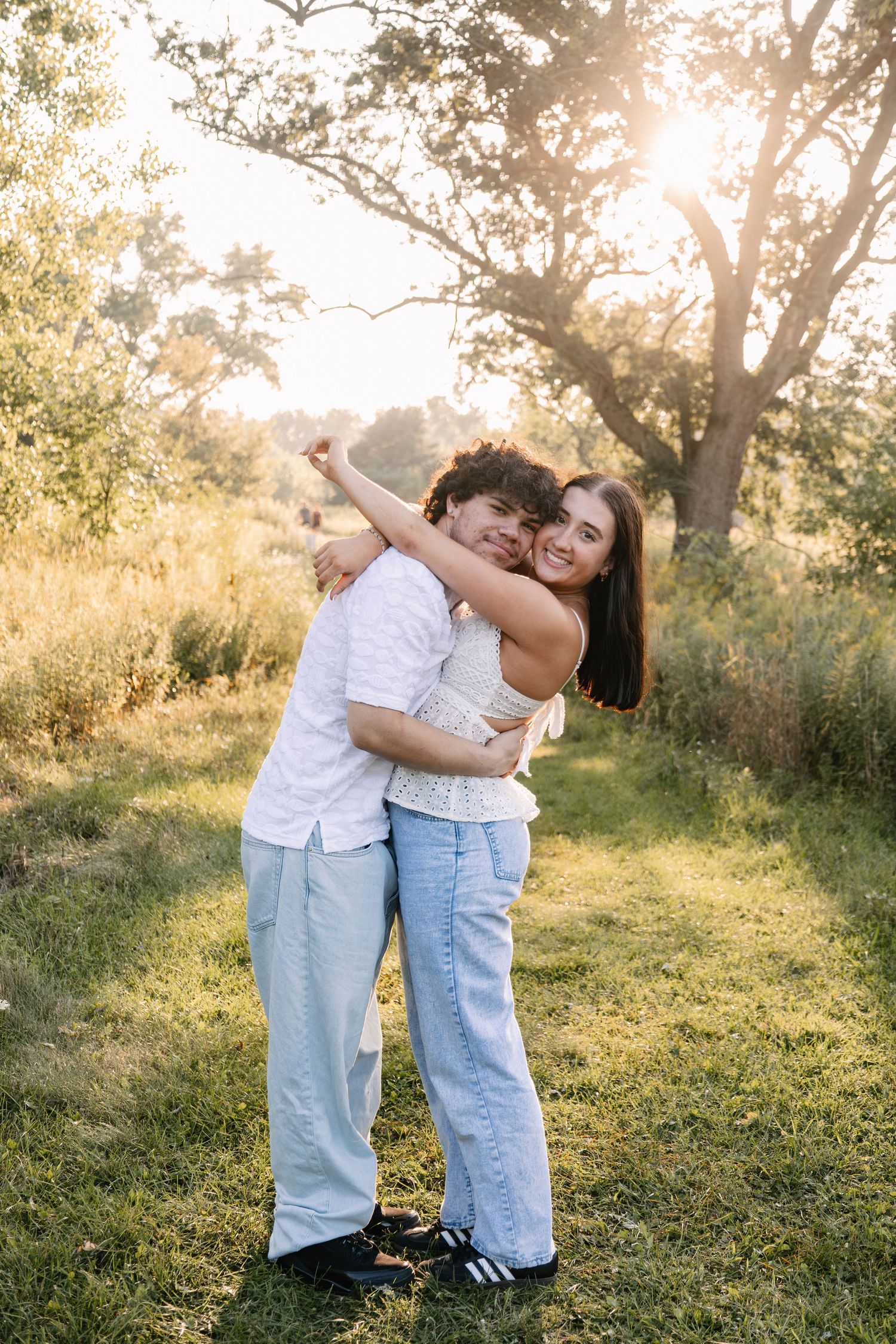 A couple embracing and smiling in a sunlit park, surrounded by lush green trees and grass during golden hour.