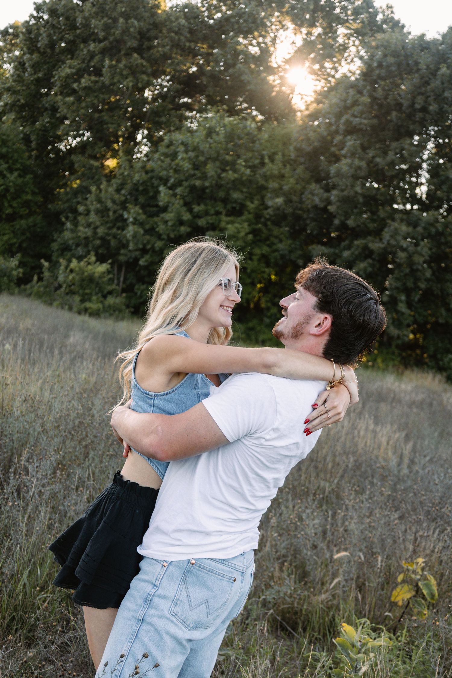 A joyful couple embraces in a sunlit meadow, smiling at each other with lush green trees in the background.