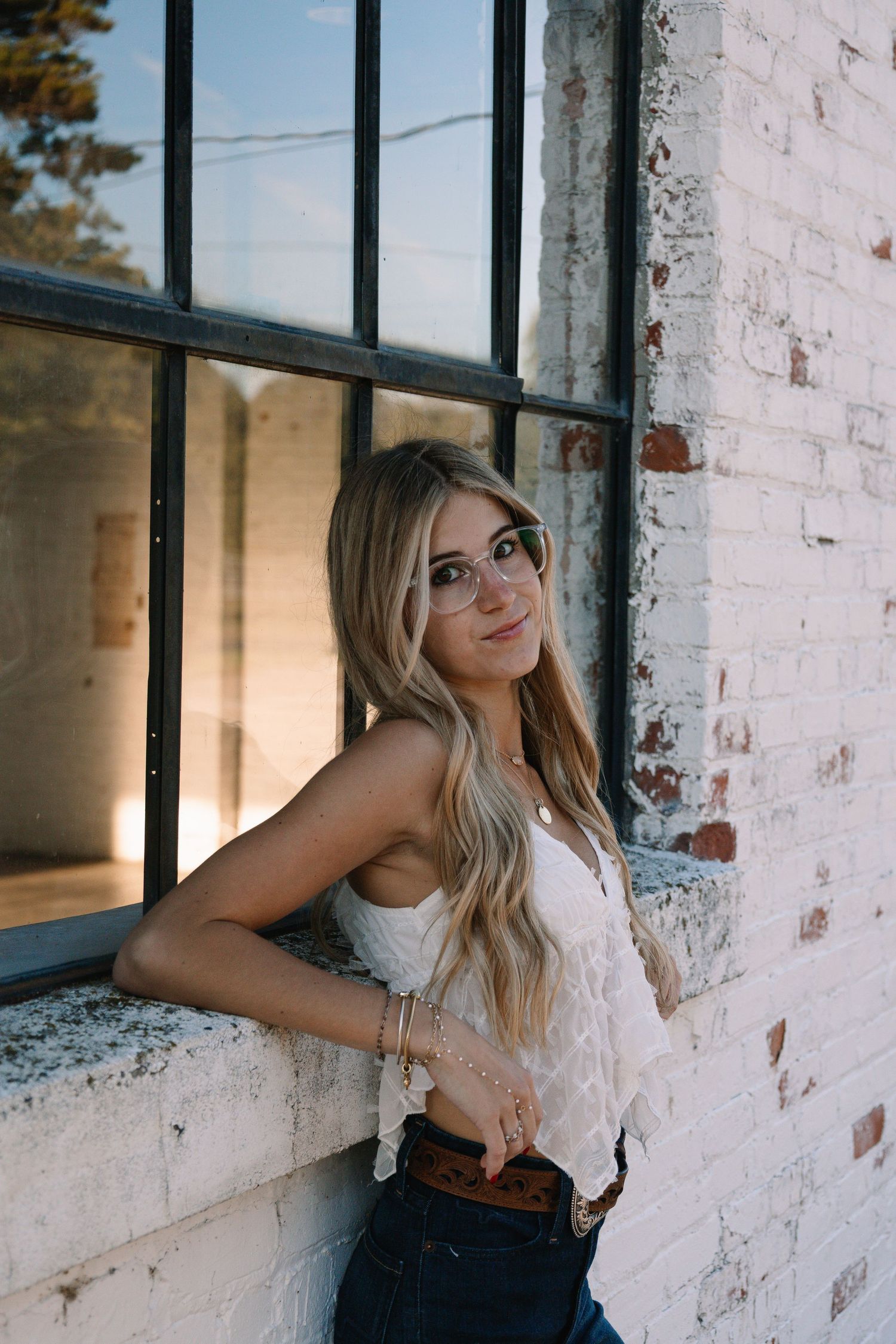 Blonde woman in white lace top leaning against whitewashed brick wall by industrial window.
