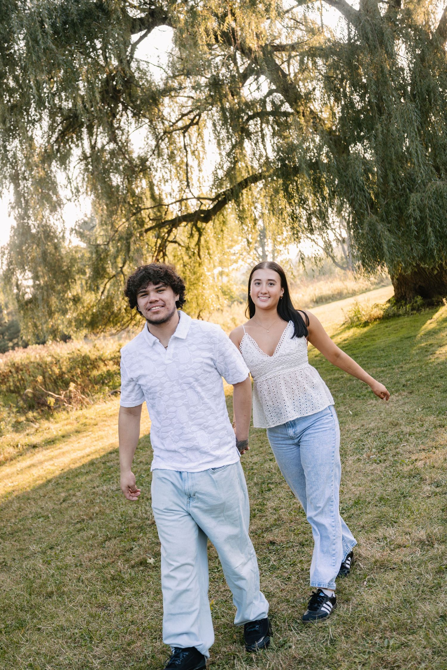 Young couple holding hands walking outdoors under a weeping willow tree at golden hour.