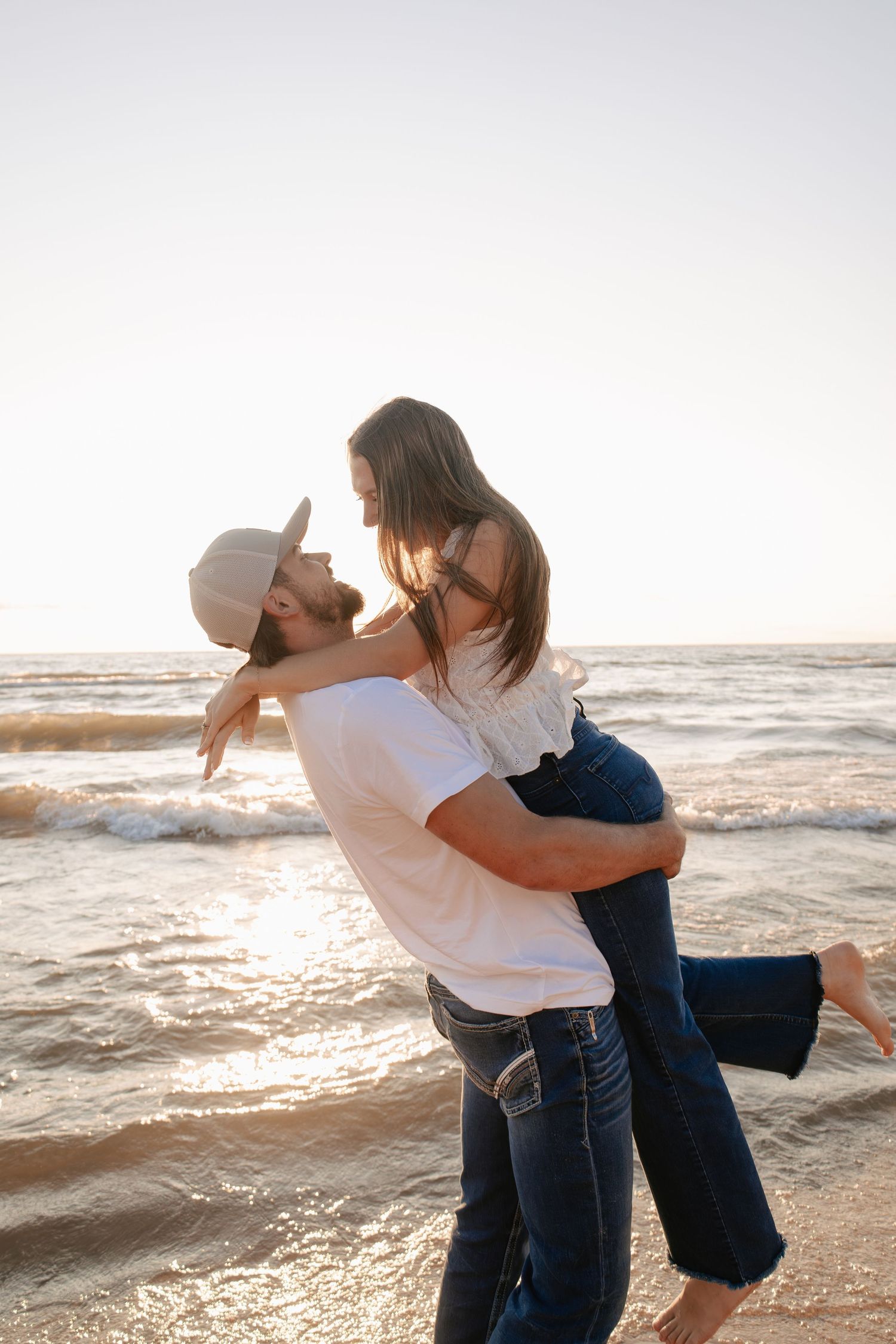 Romantic couple embracing on a sunny beach at sunset, man lifting woman in white top and jeans near ocean waves.