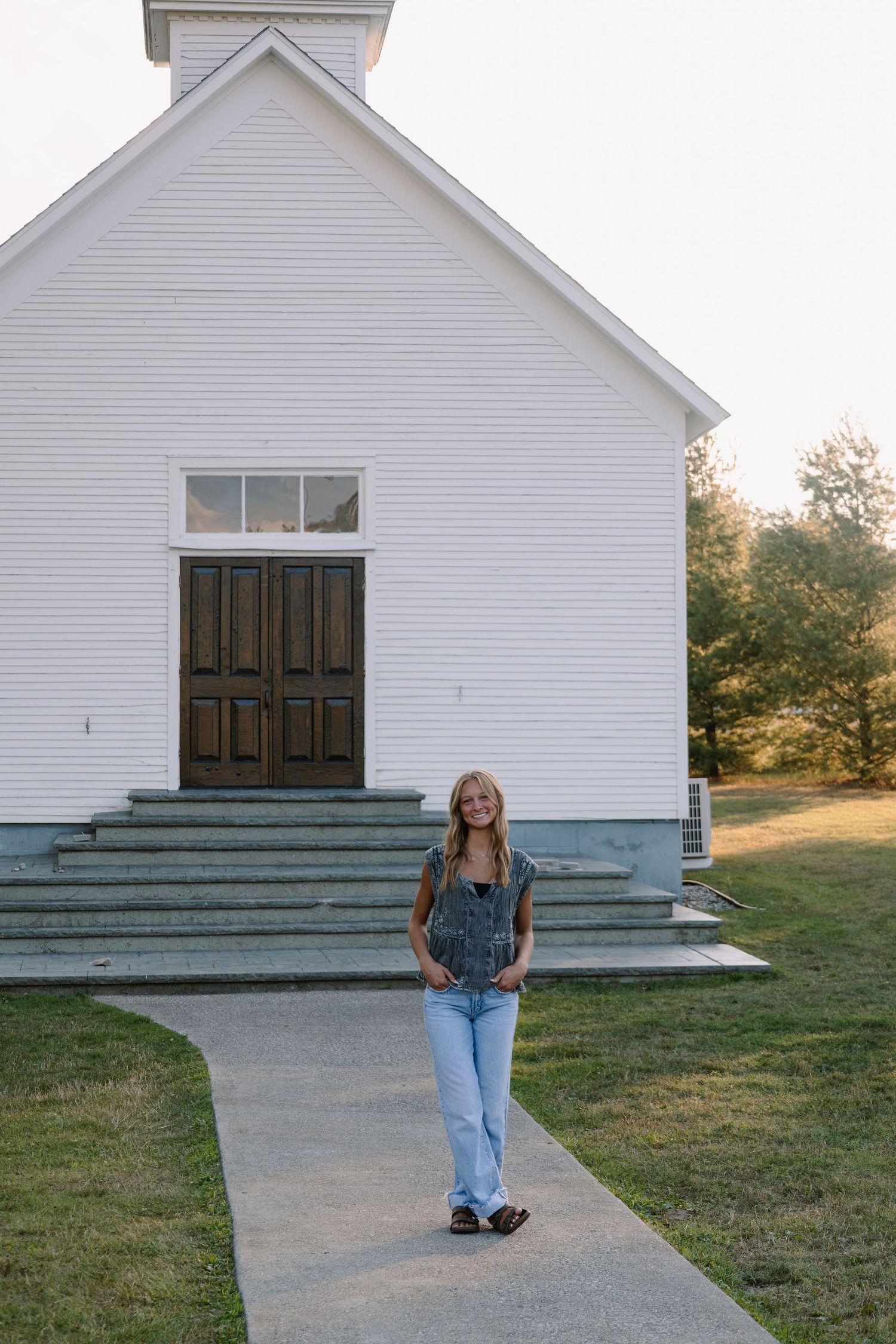 Young woman in overalls smiling on a walkway in front of a white wooden church on a sunny day.