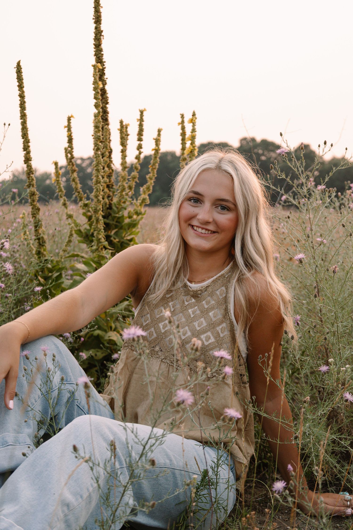 Smiling blonde woman sitting in a wildflower field wearing a tan tank top and jeans on a sunny day.