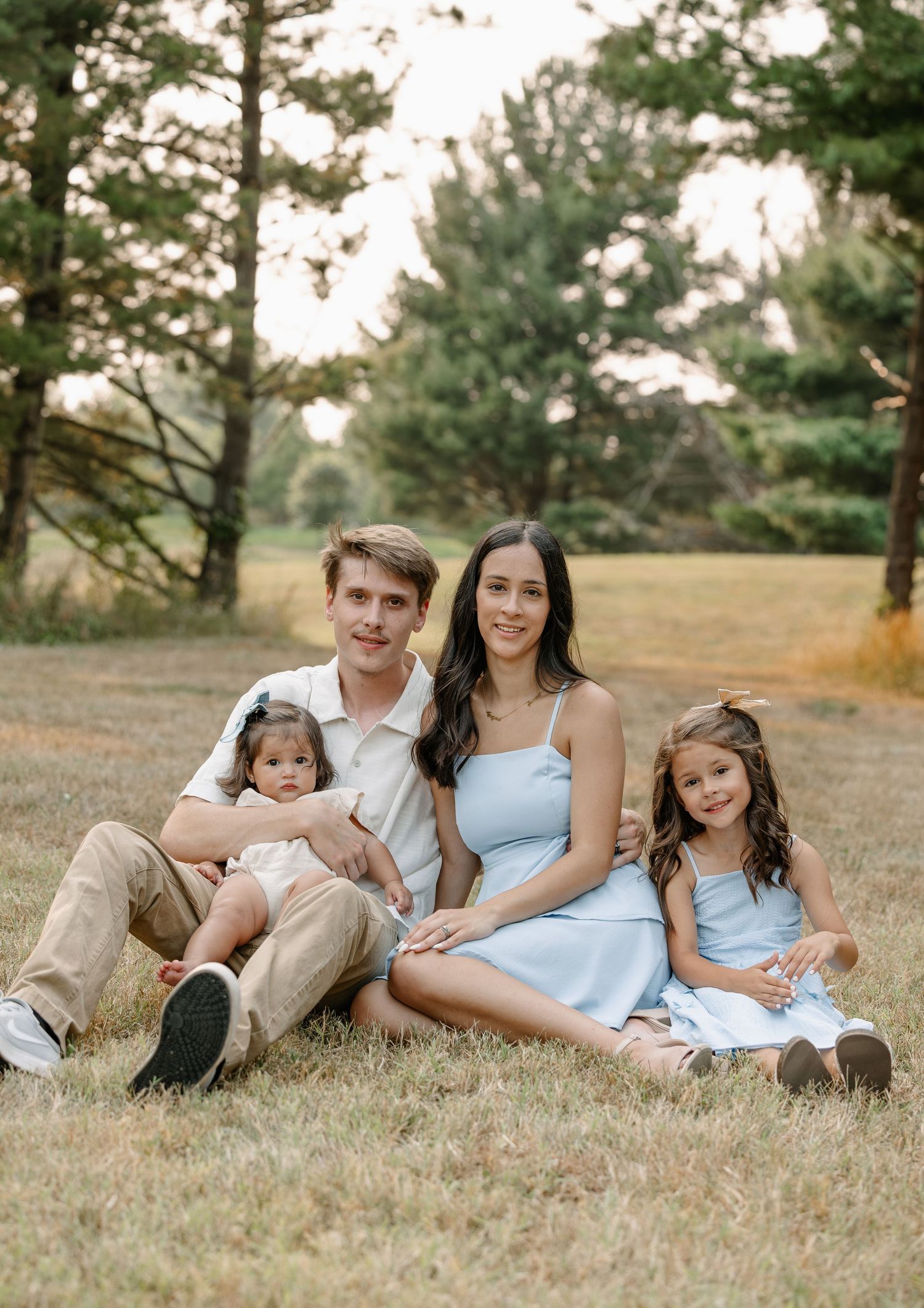 Family of four posing outdoors on grass, parents with two young daughters in light blue dresses, surrounded by trees.