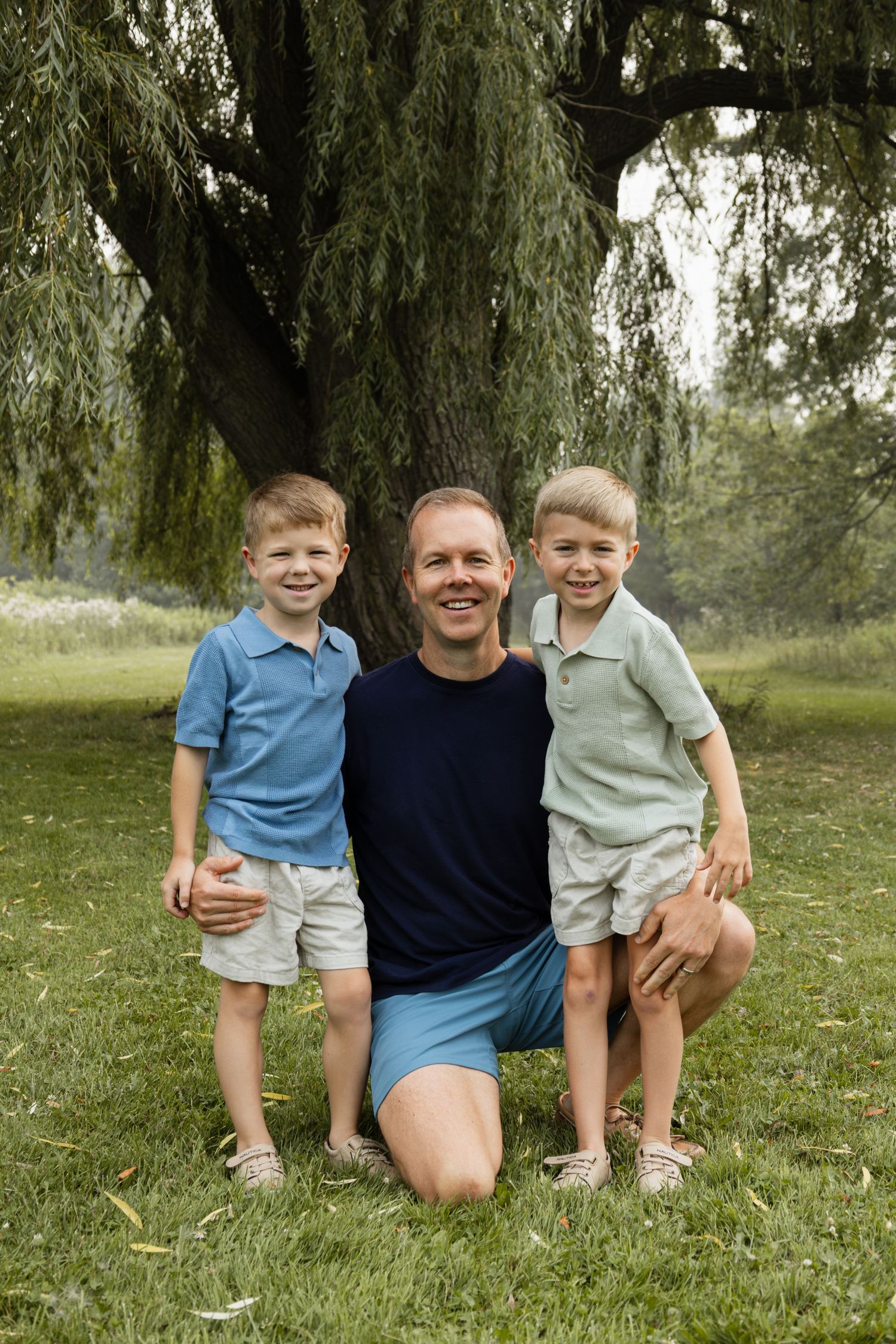 A smiling man kneels on green grass with two young blonde boys beside a willow tree outdoors.