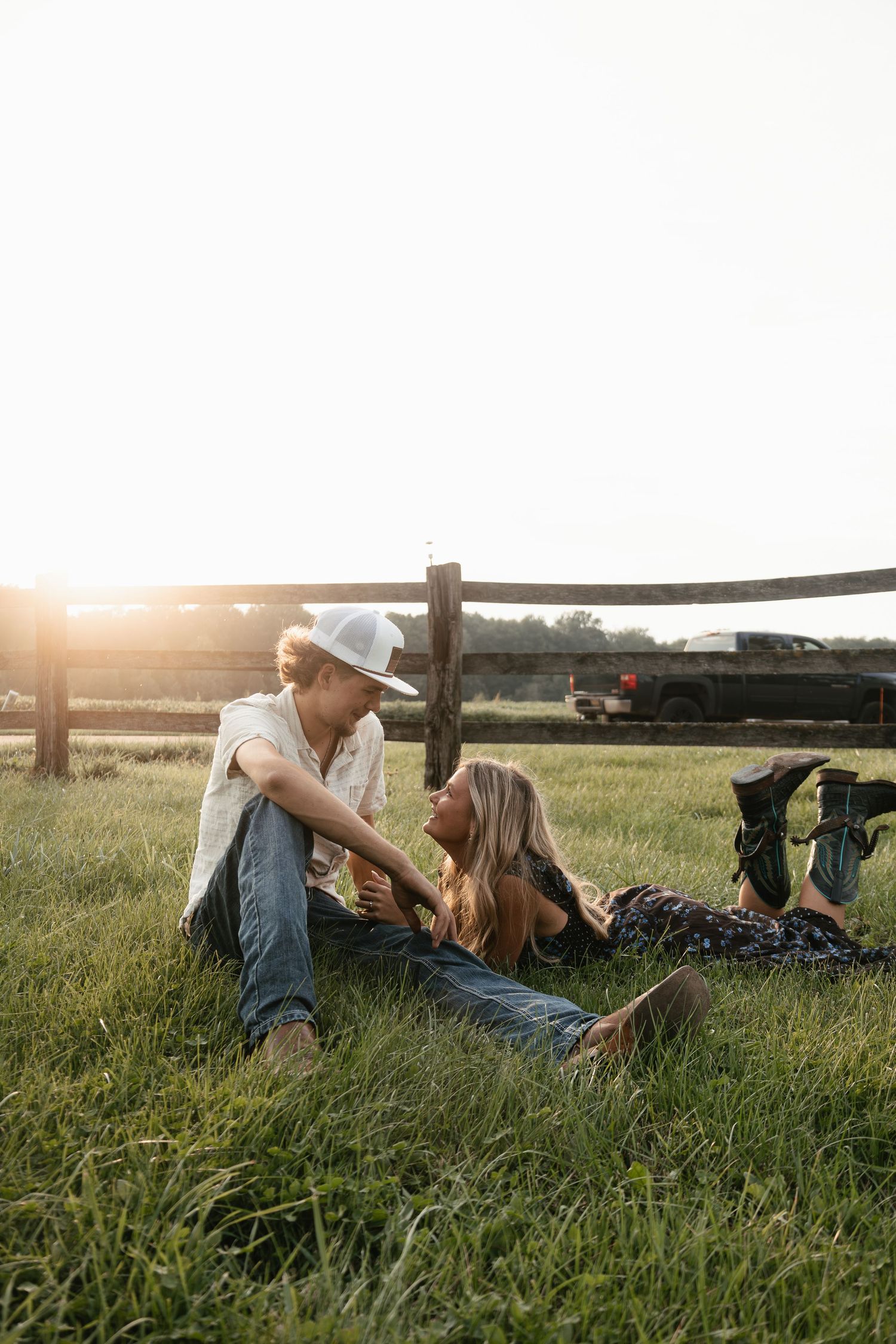 A couple sits together in a grassy field by a wooden fence at golden hour on a farm.