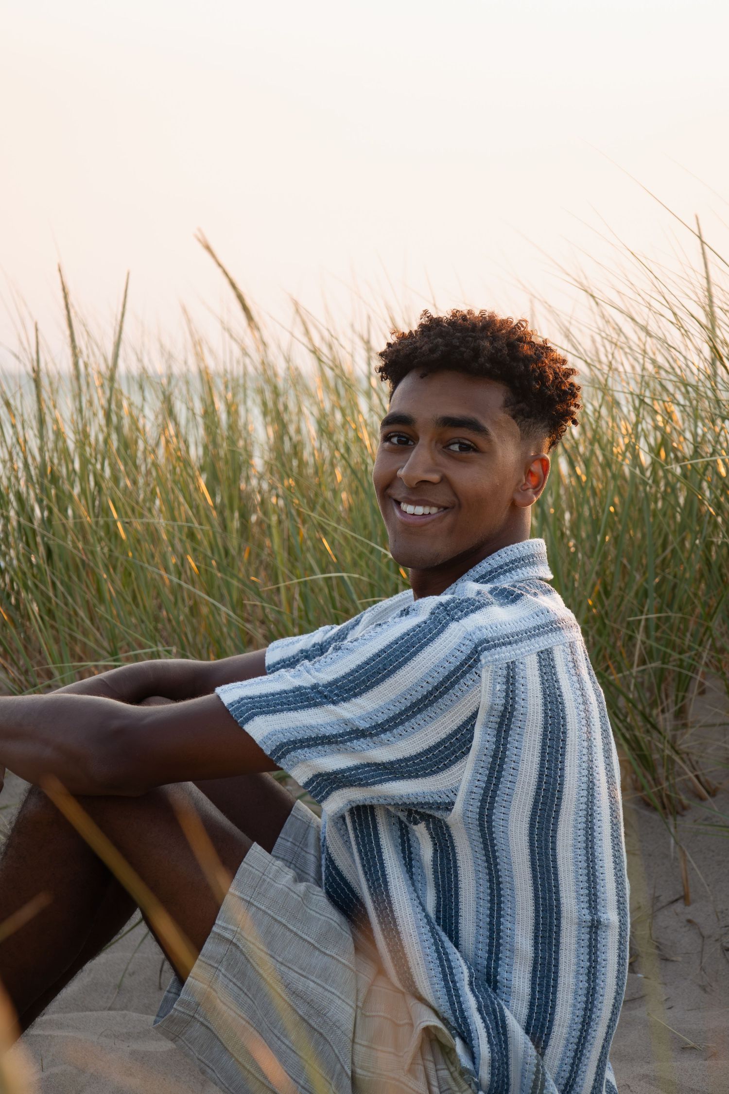 Young man in striped shirt smiling on sandy beach dunes with tall grass at sunset.