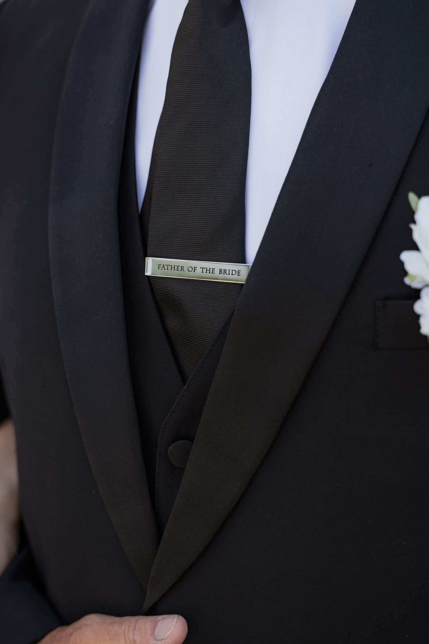 Close-up of a groom wearing a black tuxedo with a tie clip engraved 'Father of the Bride' and white boutonniere.