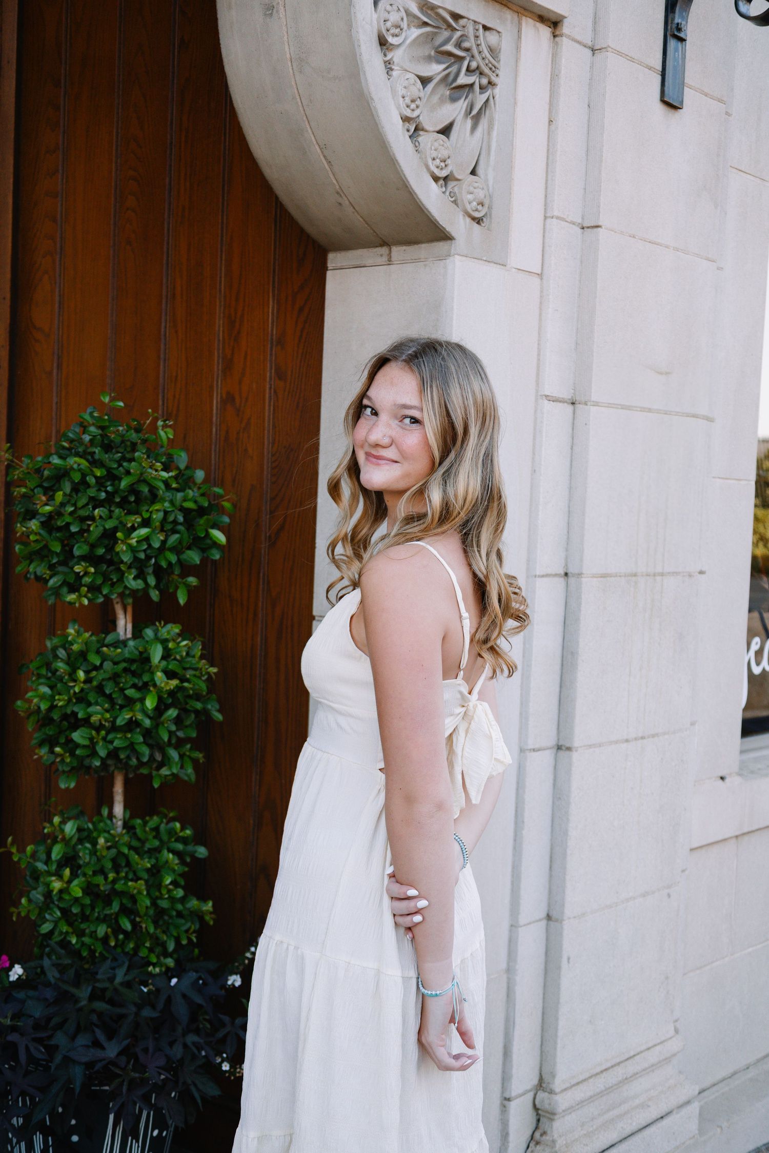 Young woman in white sundress posing elegantly by ornate stone building entrance with topiary bushes.