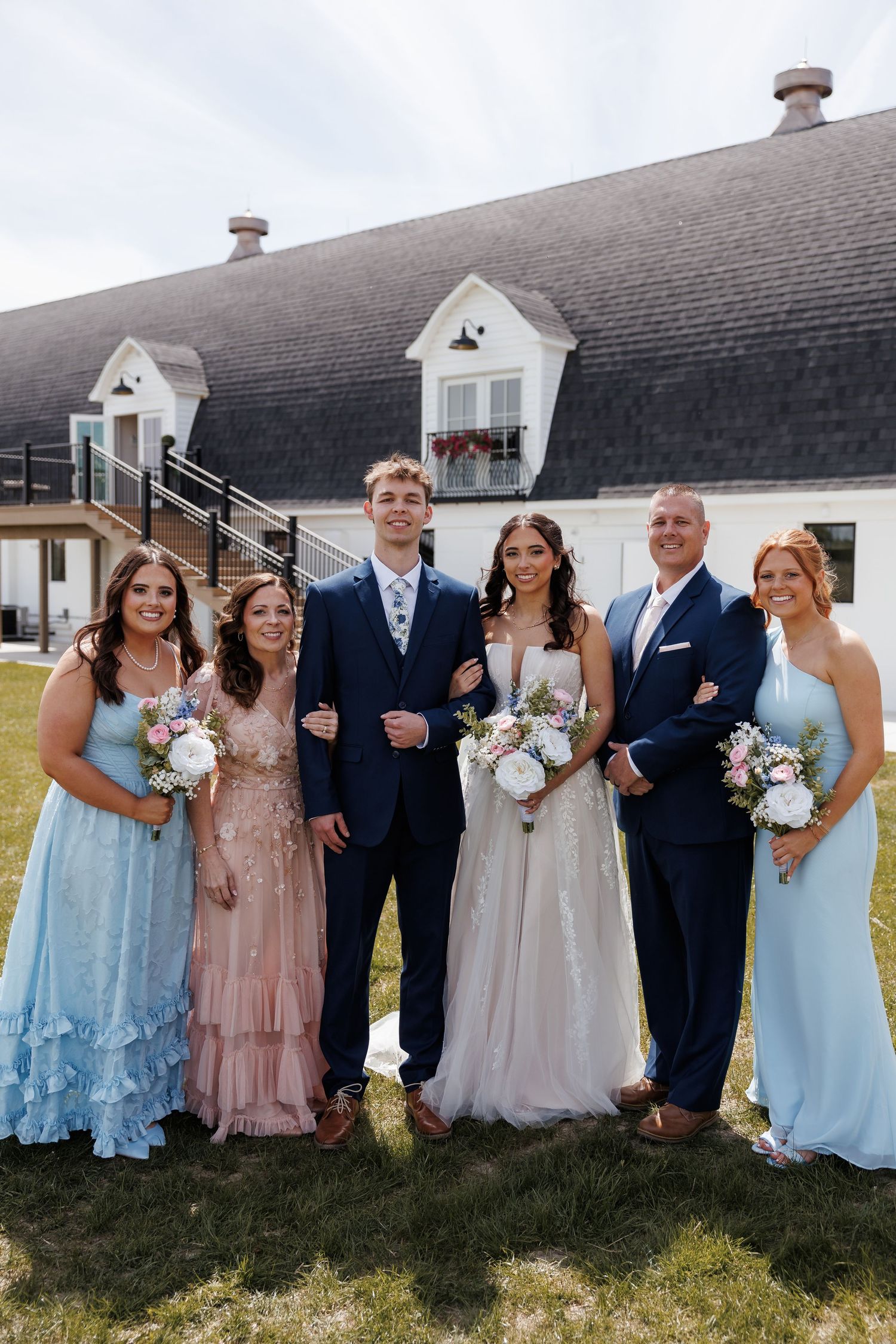 Wedding party family portrait with bride, groom, and four guests outside white barn venue.