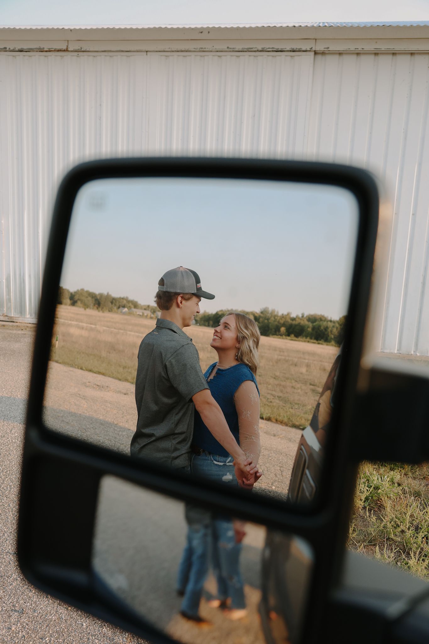 A couple holds hands and gazes at each other, reflected in a truck side mirror with an open field in the background.