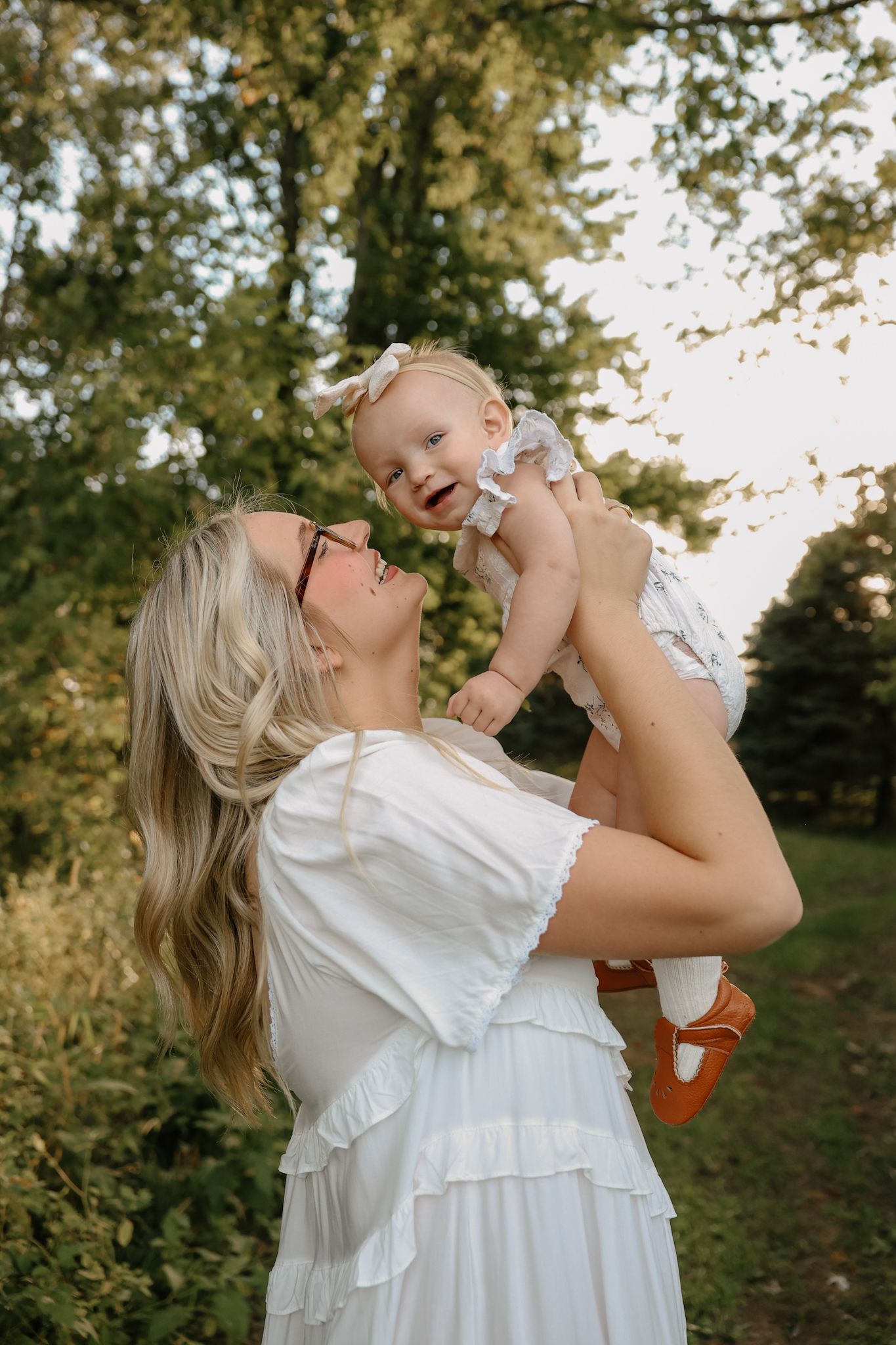 Joyful mother in white dress lifting laughing baby girl in white romper in sunlit outdoor setting.