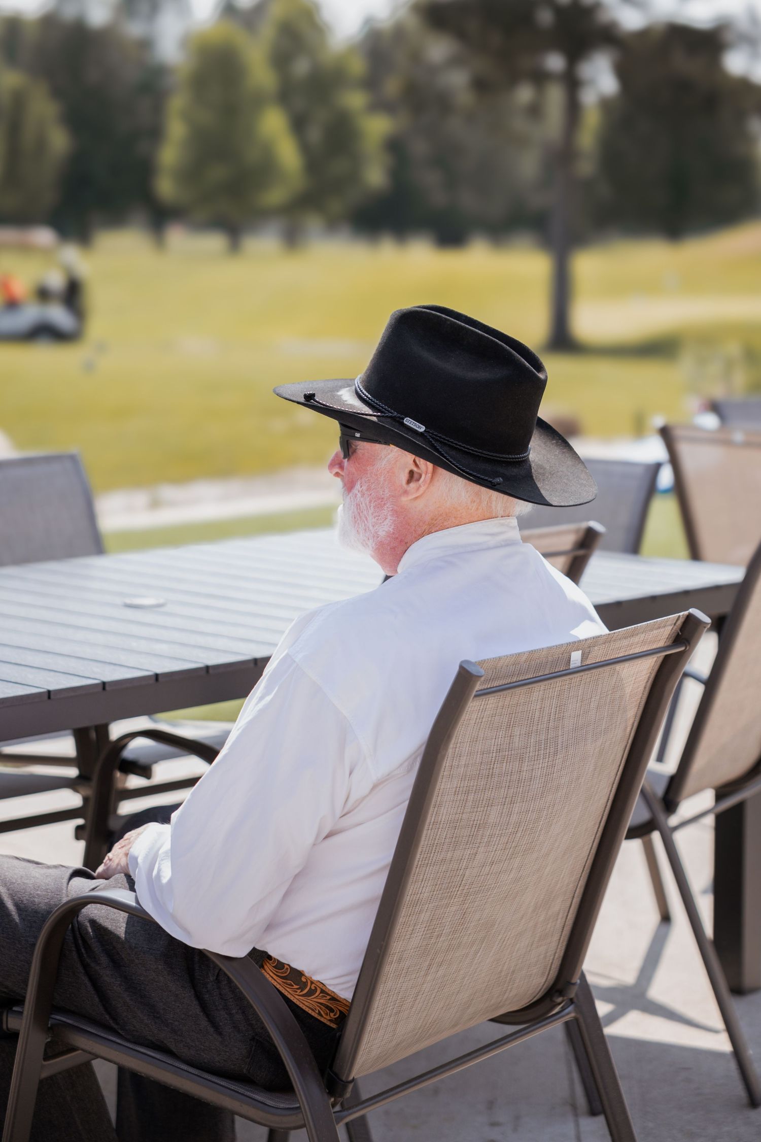 Elderly man in black cowboy hat and white shirt seated at outdoor patio table at wedding.