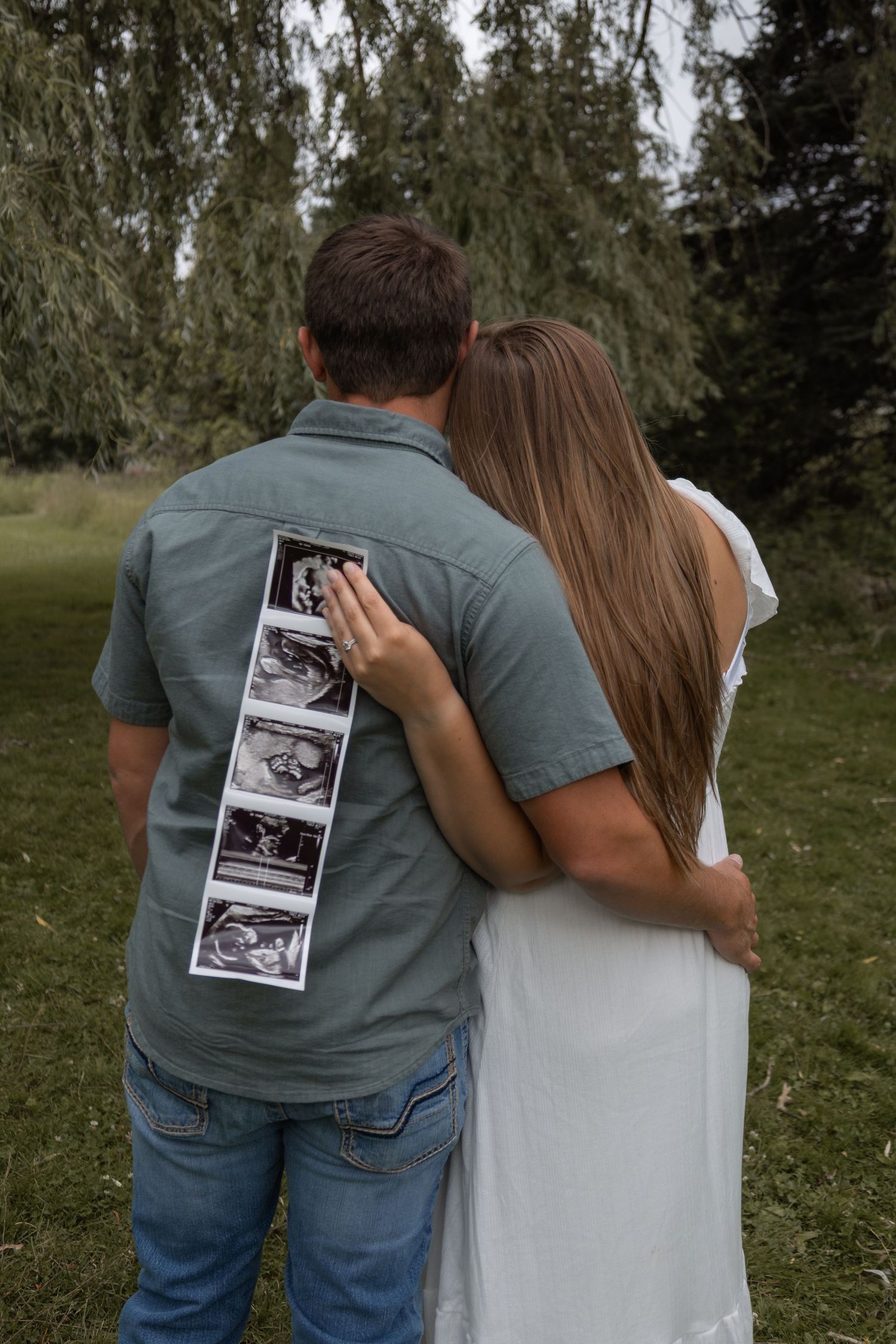 Couple standing back to back with woman displaying ultrasound strip photos on mans back under willow tree.