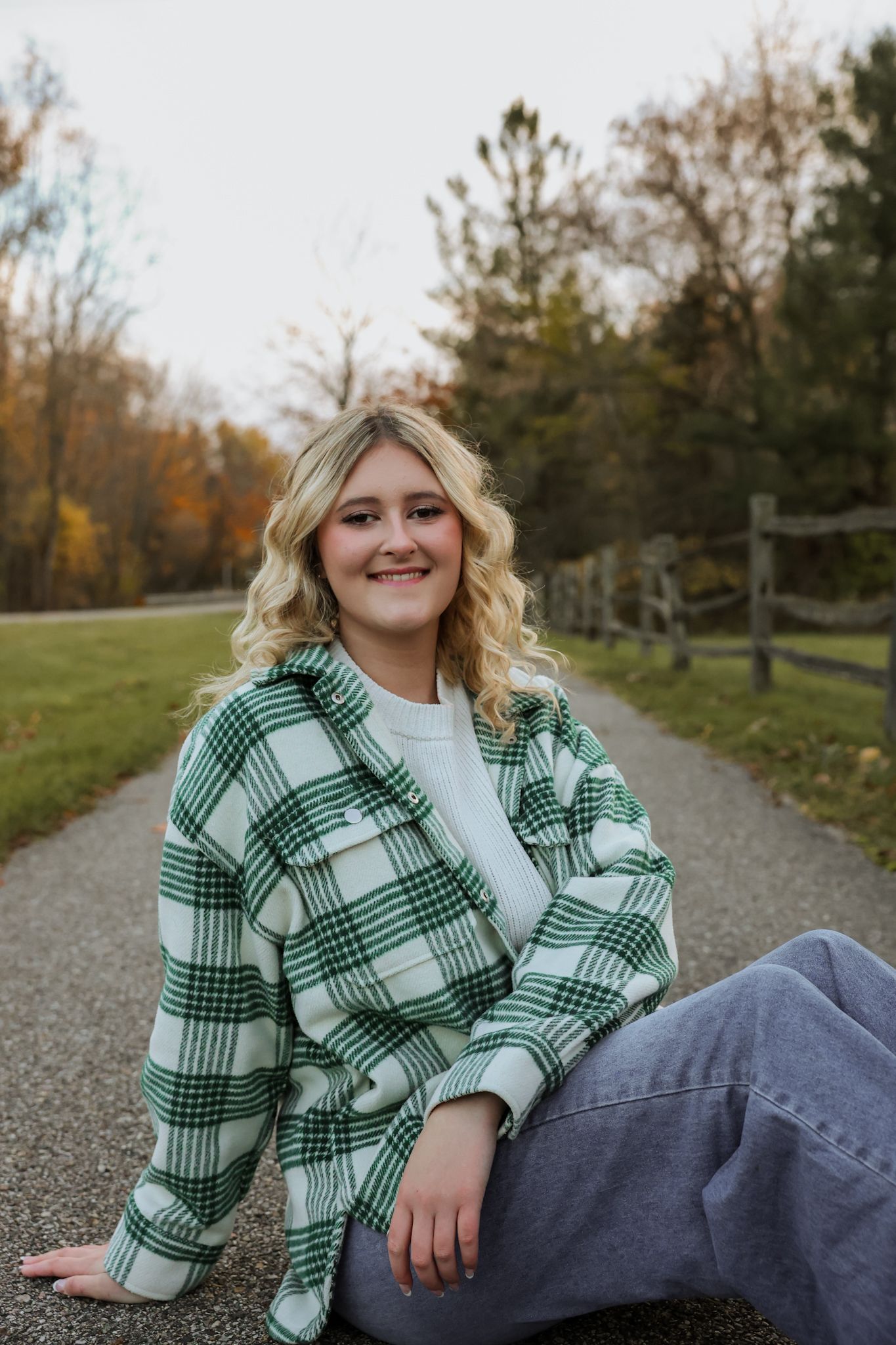 Smiling blonde teen in green plaid jacket sitting on pathway surrounded by autumn trees.