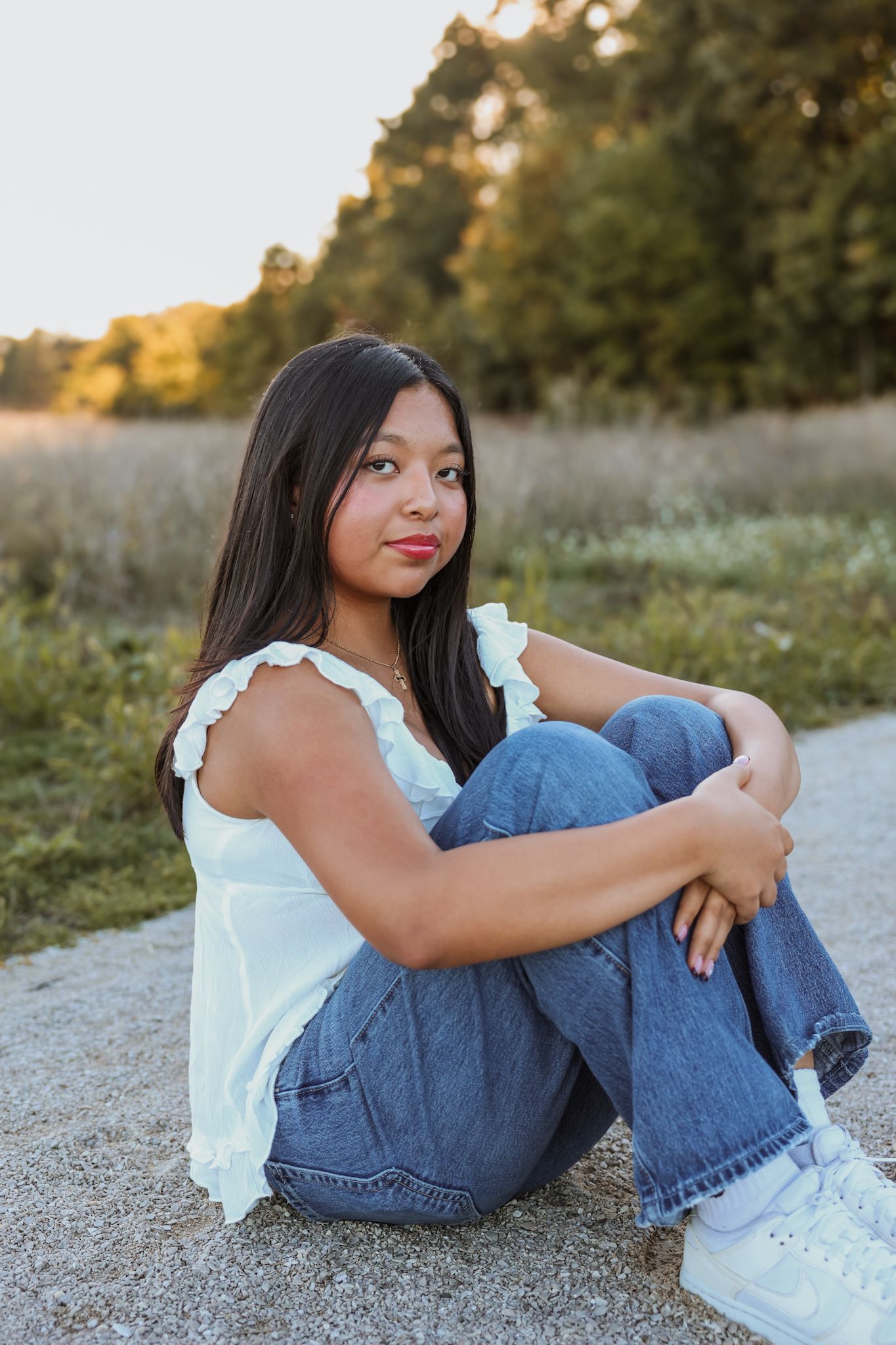 Young Asian woman in white top and jeans sits relaxed on a gravel path at golden hour.