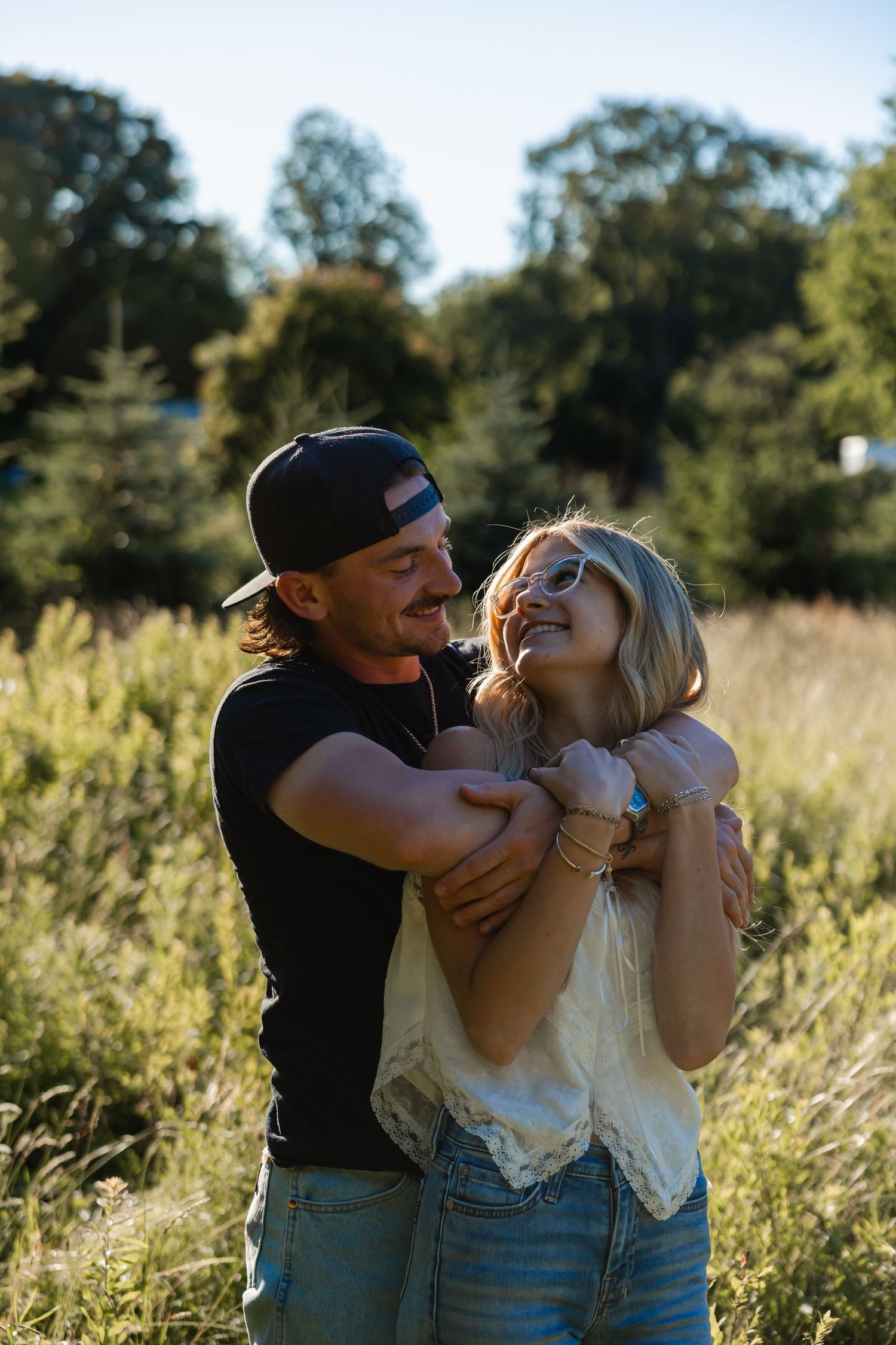 Man in black cap embracing woman from behind in a sunny meadow, both laughing joyfully outdoors.