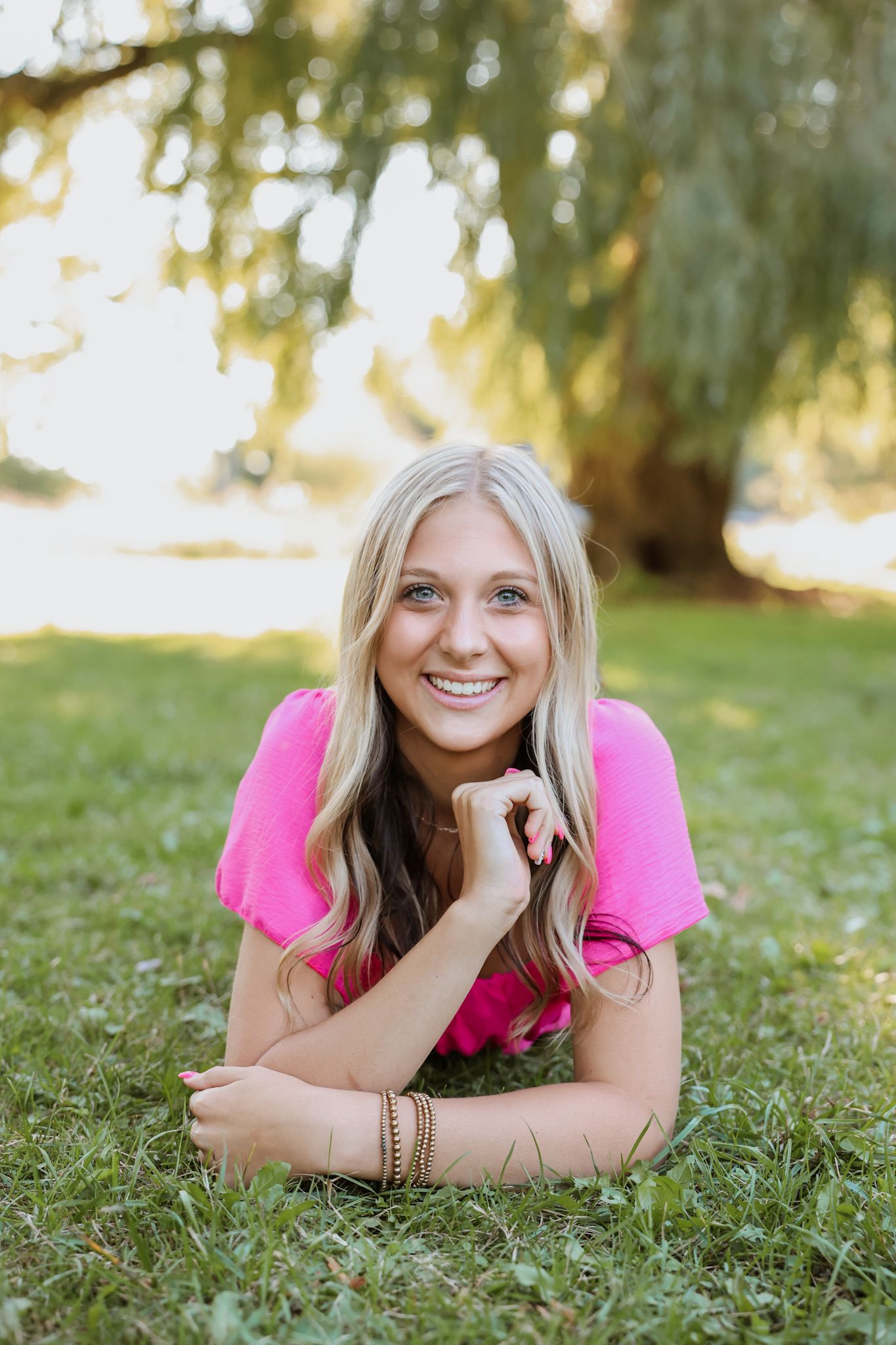 Smiling blonde woman in pink top lying on green grass under a willow tree in soft sunlight.