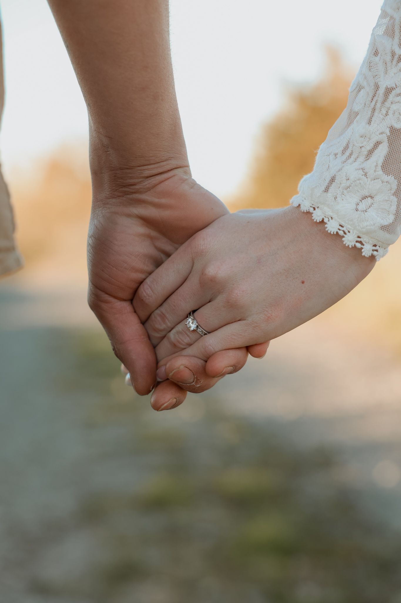 Couple holding hands at sunset, showcasing a diamond engagement ring on the woman's finger.