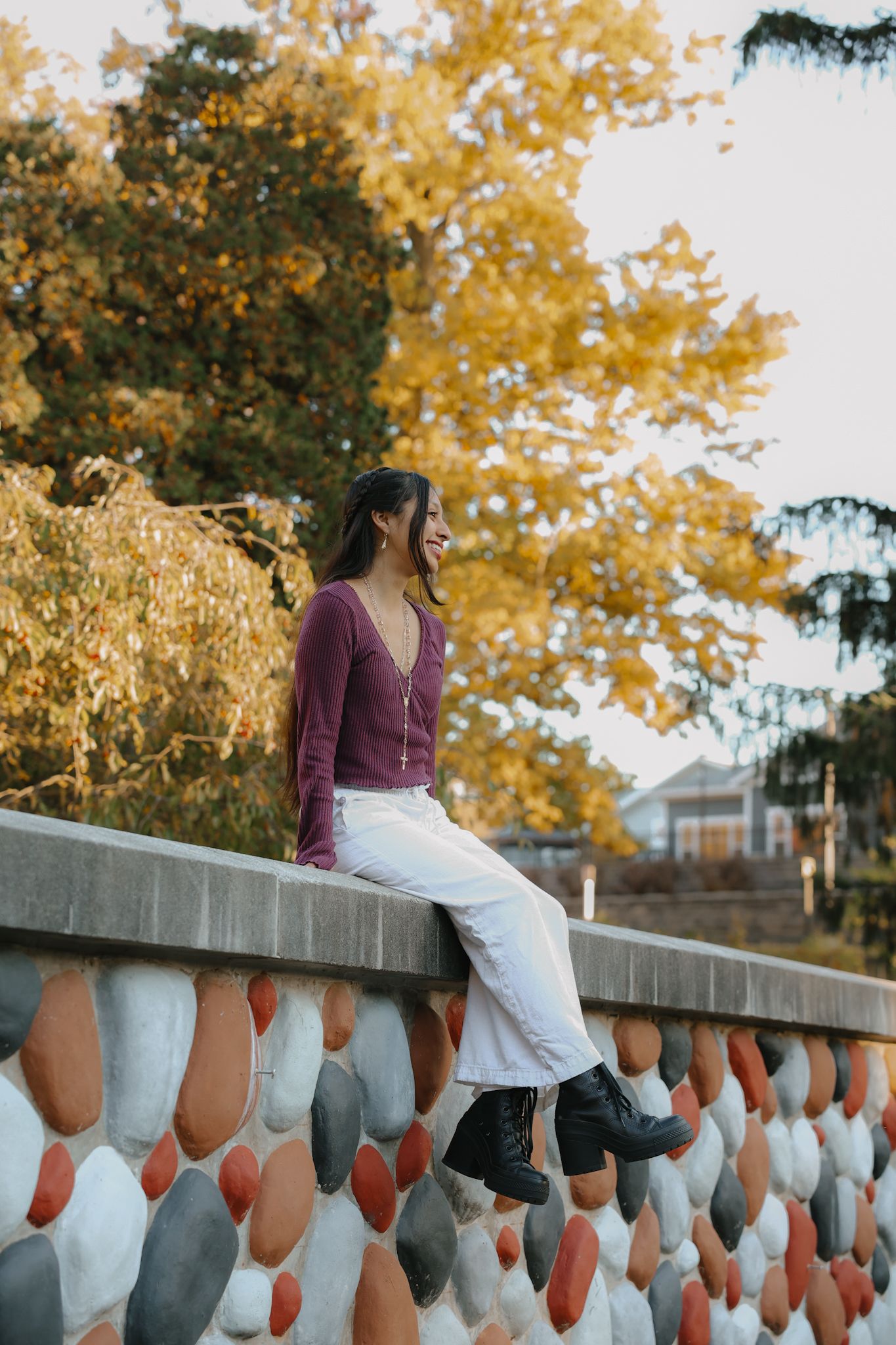 Young woman in purple top and white pants sitting on decorative stone wall with fall foliage.