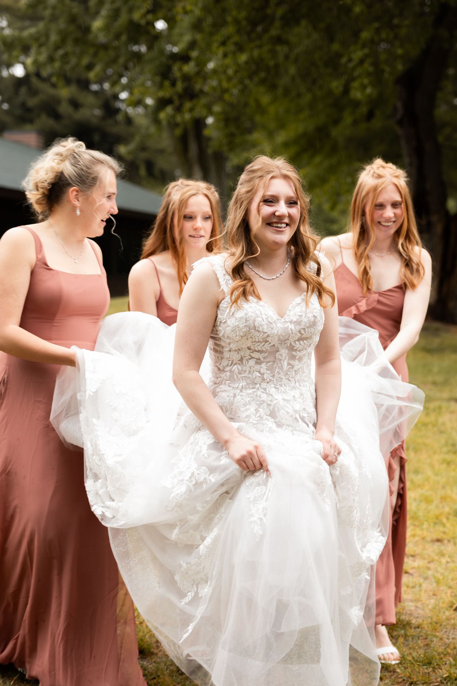 Bride in lace gown walks outdoors with three bridesmaids in dusty rose dresses.