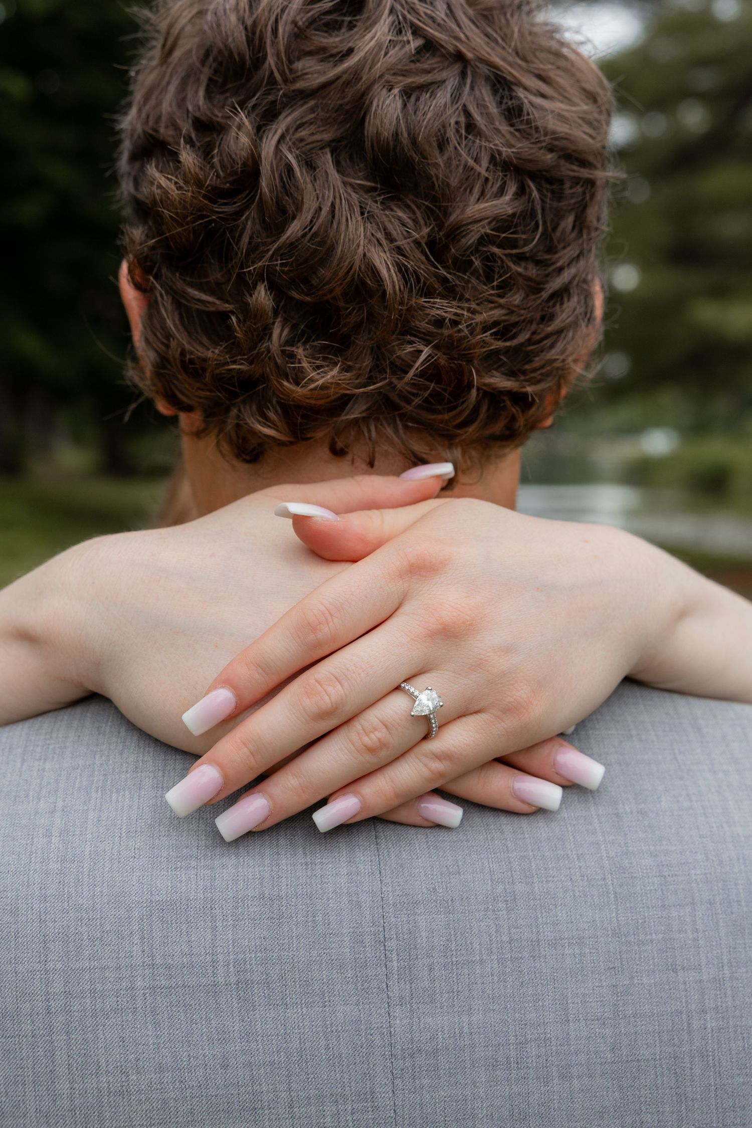 Close-up of bride's hand with diamond engagement ring resting on groom's gray suit jacket.
