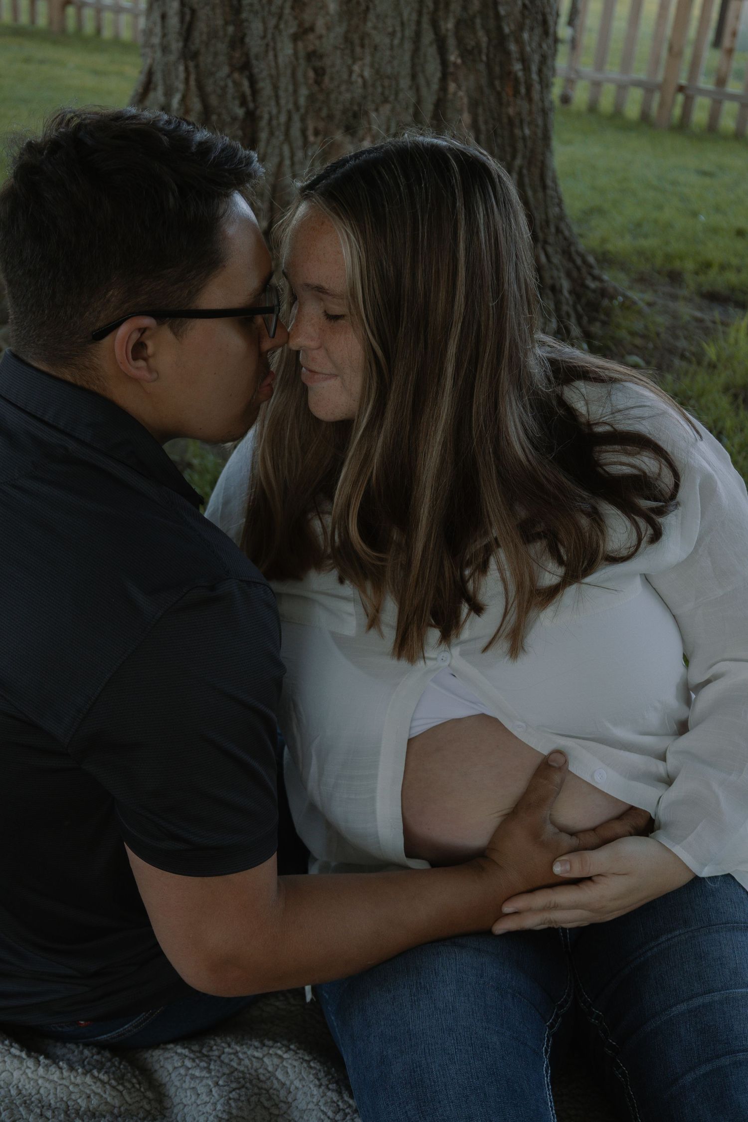 Couple sharing tender moment under tree, man kissing pregnant woman's cheek while holding her baby bump.