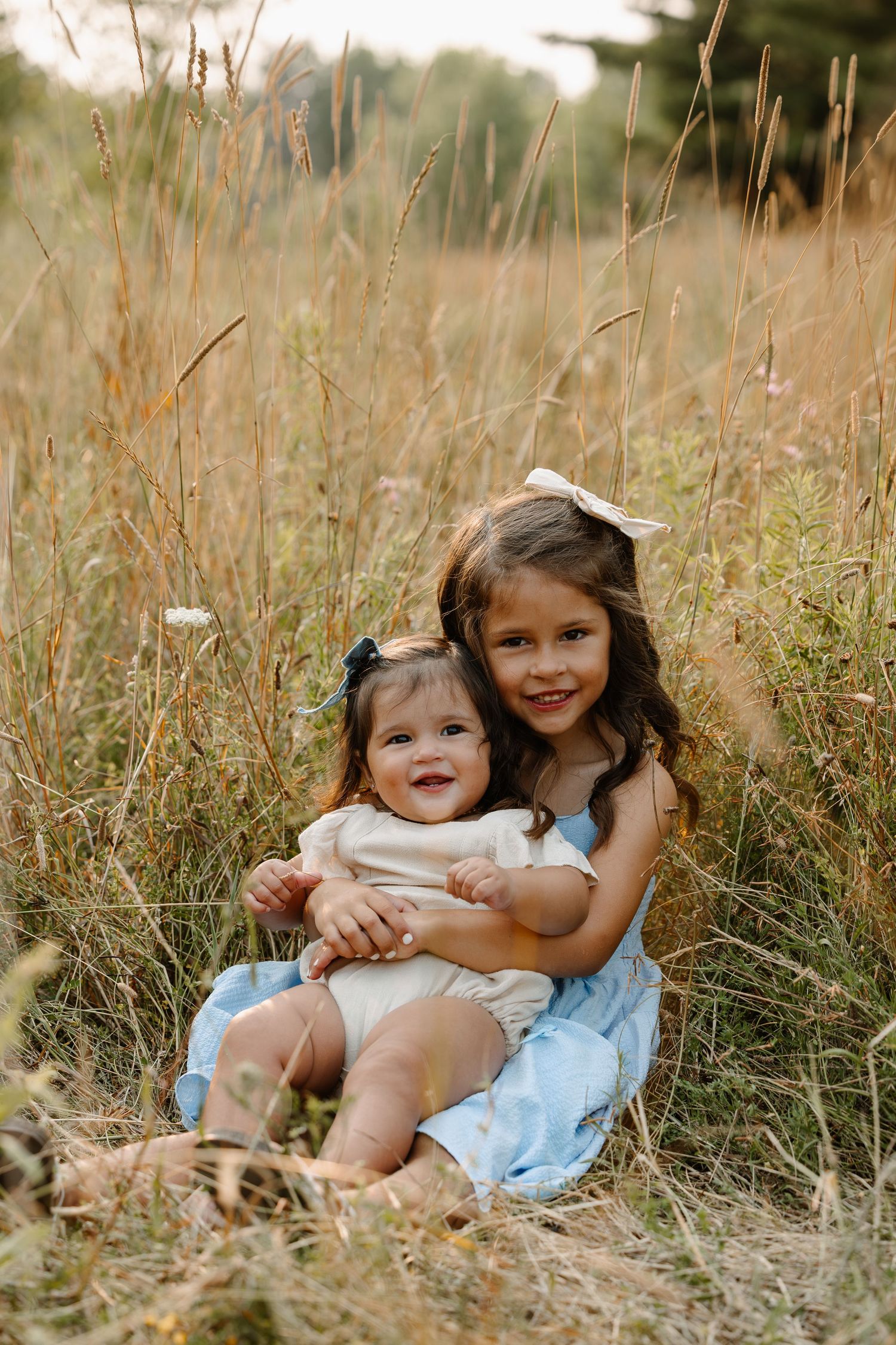 Two young sisters sitting in a golden field, older girl in blue dress hugging smiling baby sister.