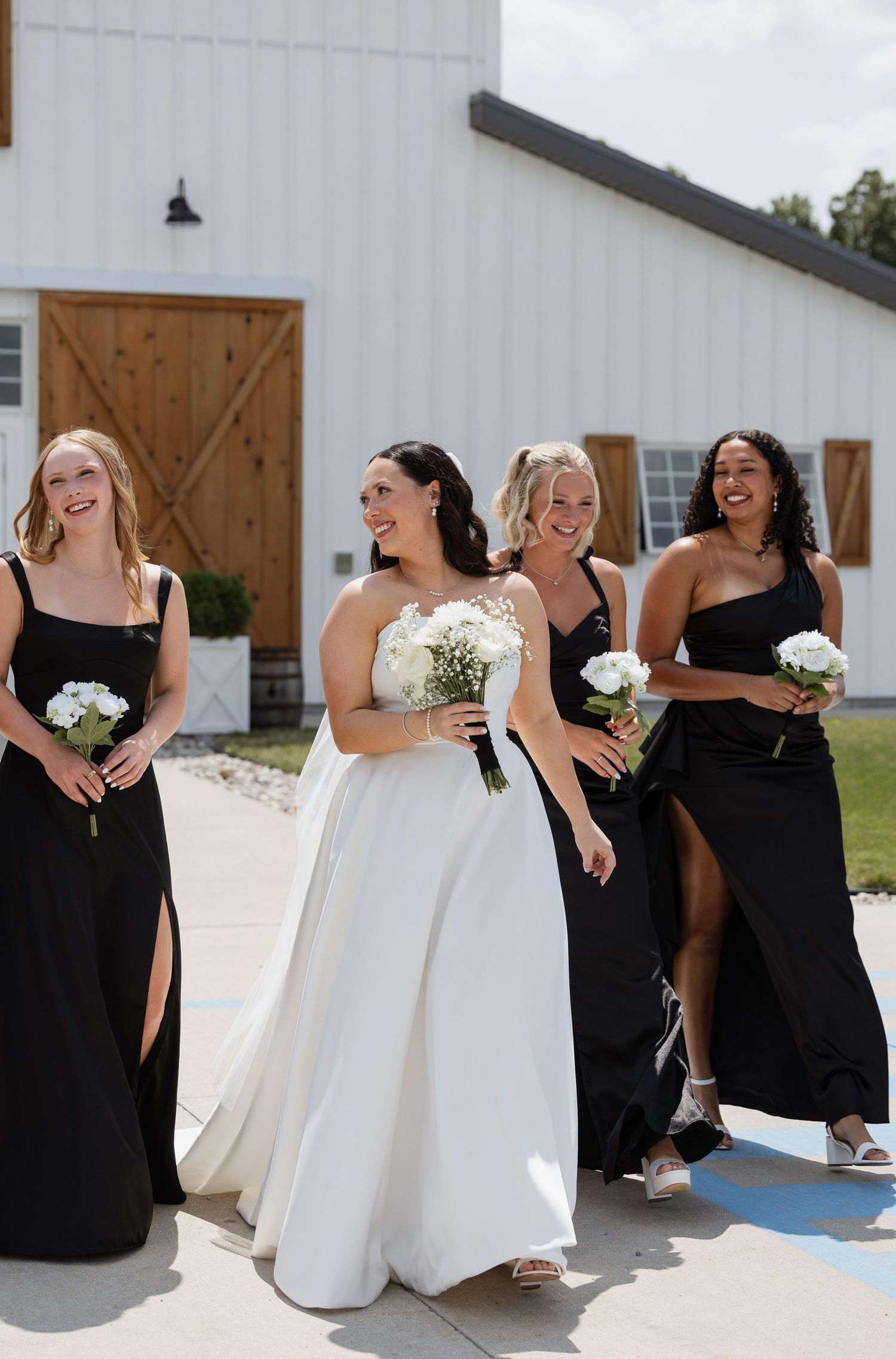 Bride in white gown walks with three bridesmaids in black dresses, holding white bouquets outside a white barn.
