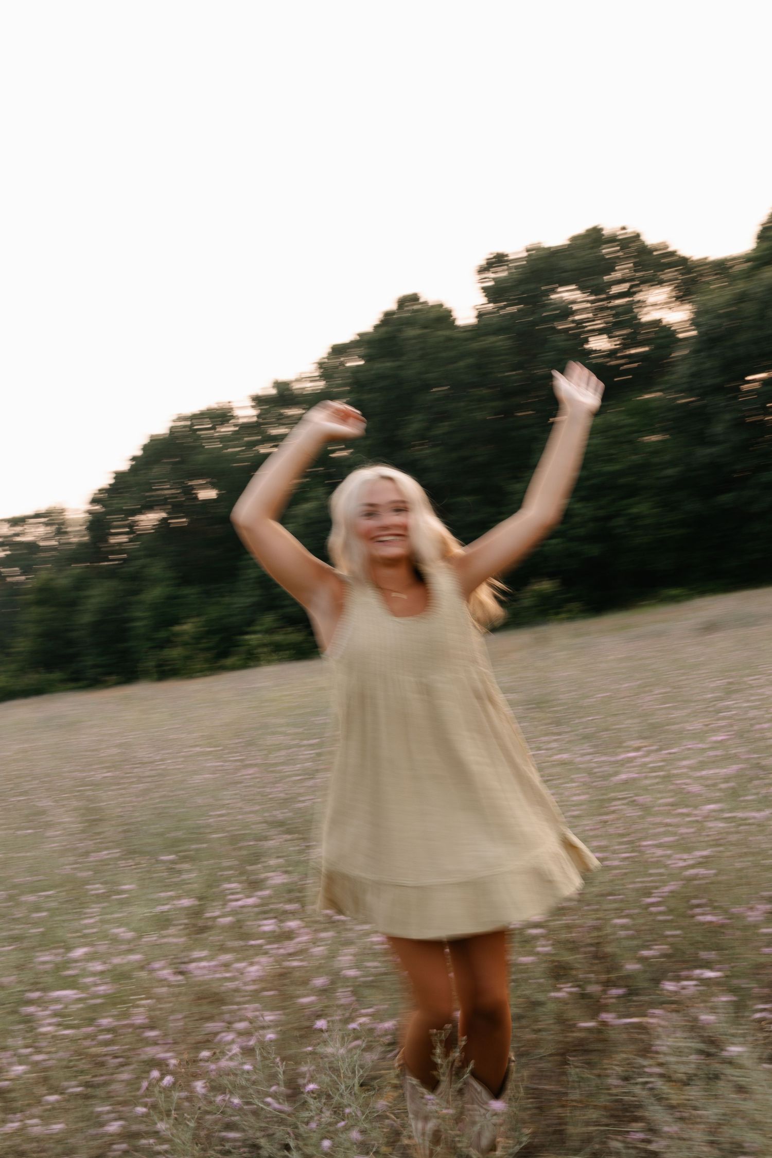 Joyful blonde woman in beige mini dress dancing in wildflower field with arms raised, surrounded by green trees.