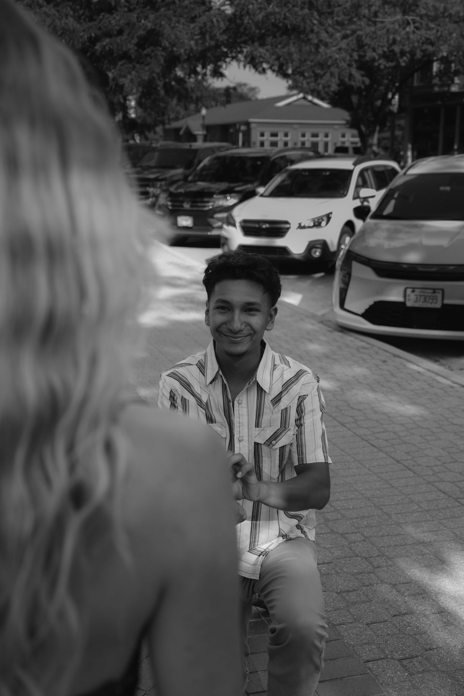 Black and white photo of a smiling young man in a striped shirt kneeling outdoors in a parking lot.