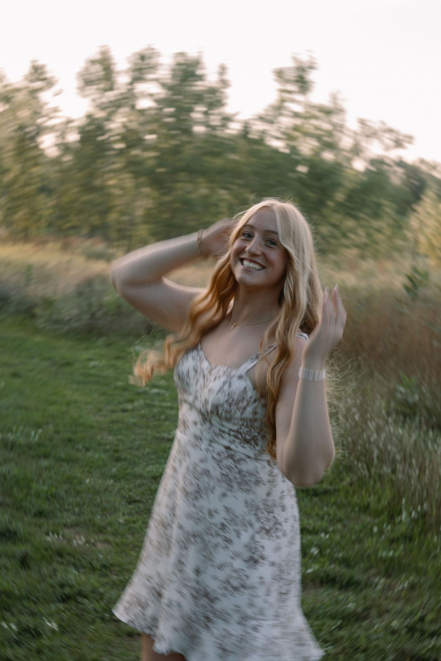 Smiling blonde woman in floral sundress twirling outdoors in lush green field at golden hour.