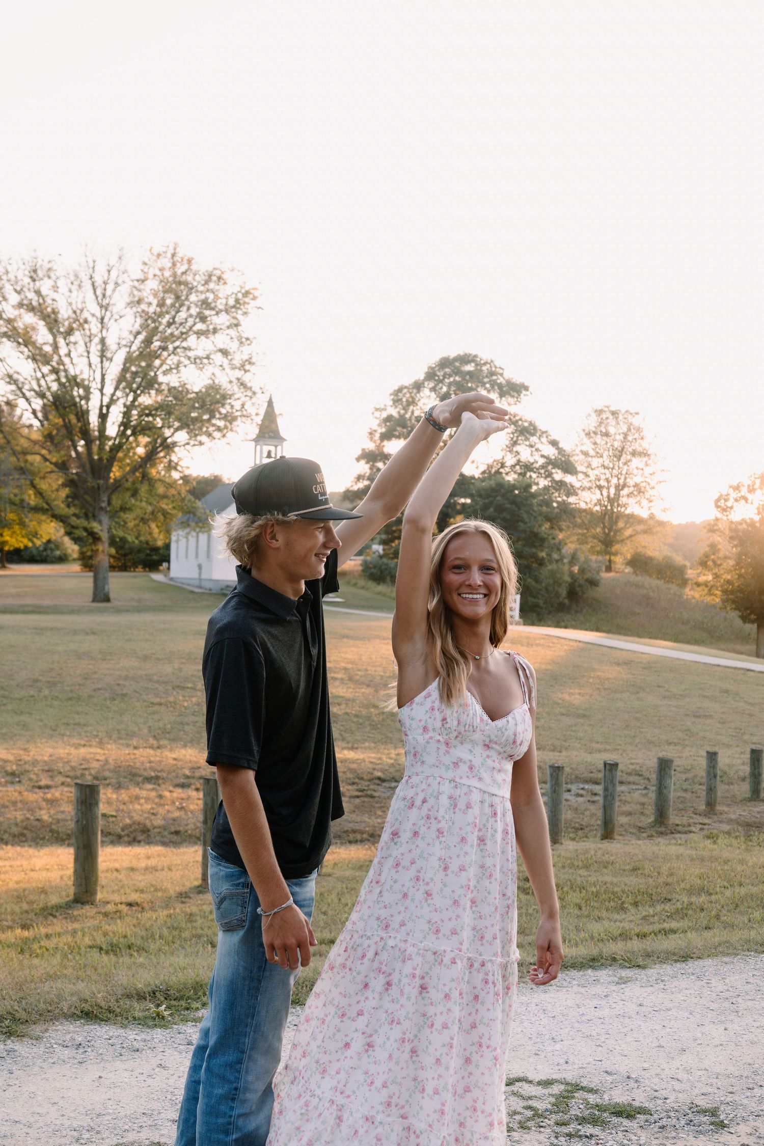 A couple dances outdoors at golden hour near a white country church, woman in floral dress twirling.