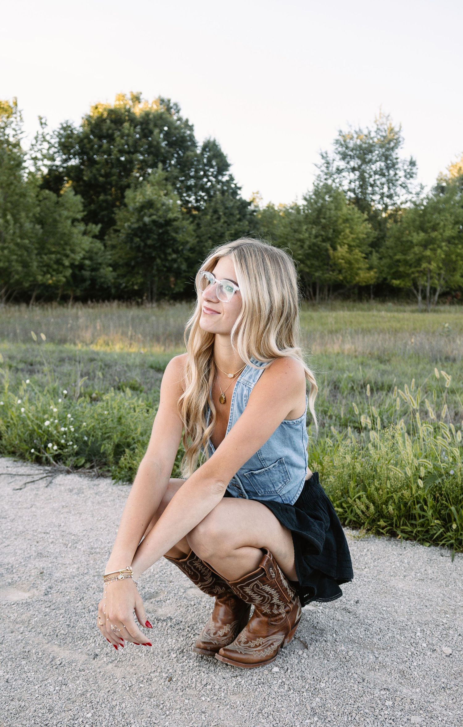 Blonde woman in denim top, black skirt, and cowboy boots crouching outdoors in a field wearing mirrored sunglasses.