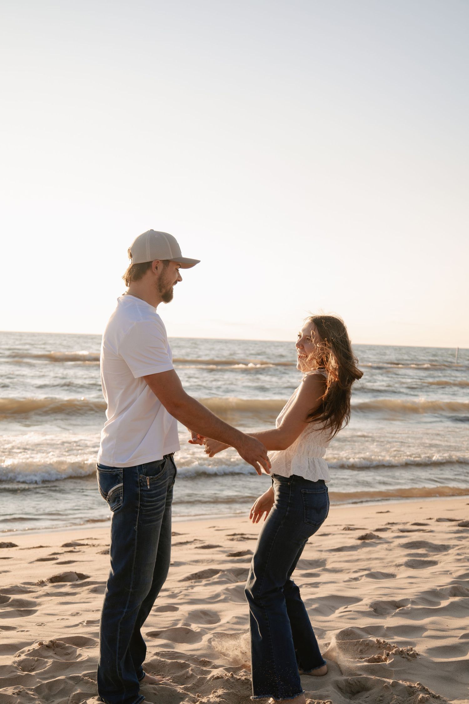 Couple holding hands and dancing on sunny beach at sunset, both wearing white tops and jeans near ocean waves.