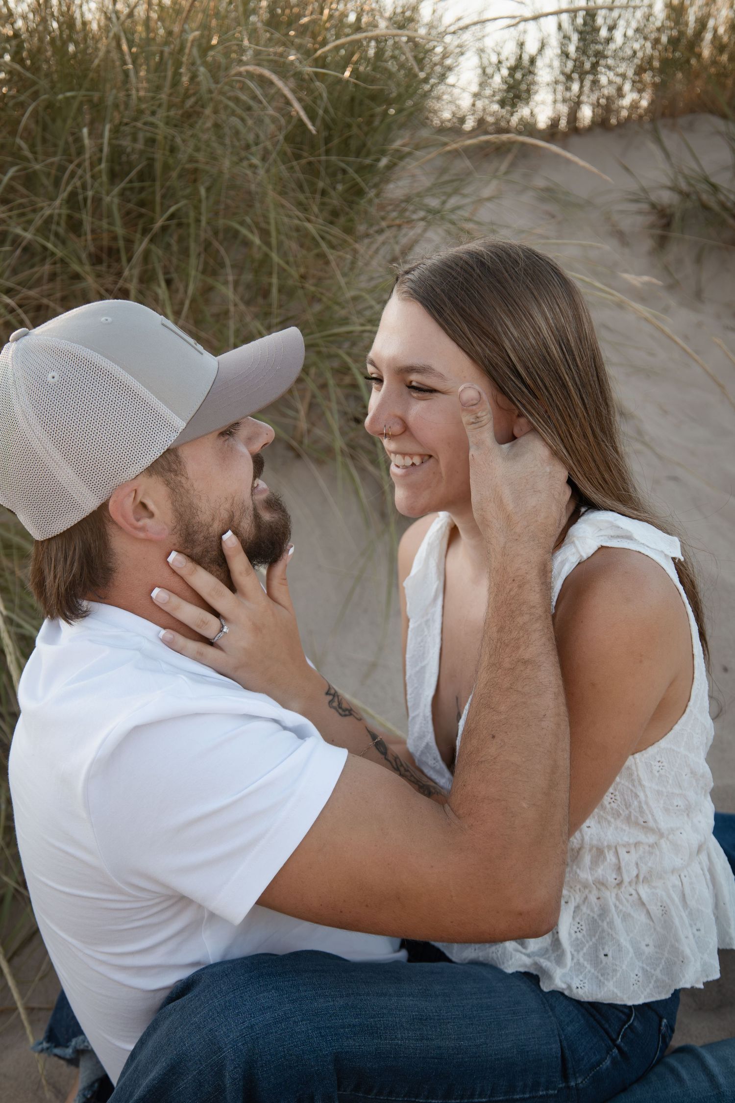 A couple shares a romantic moment on sandy beach dunes, smiling at each other tenderly.