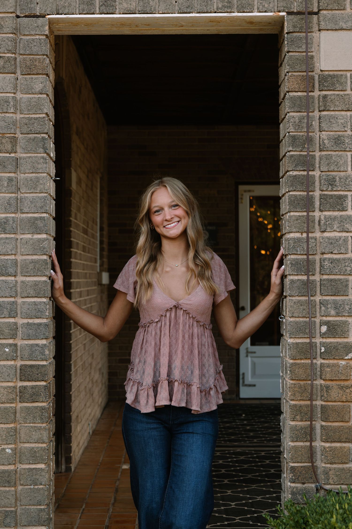 Smiling young woman in pink ruffled top and jeans posing in brick doorway of a home.