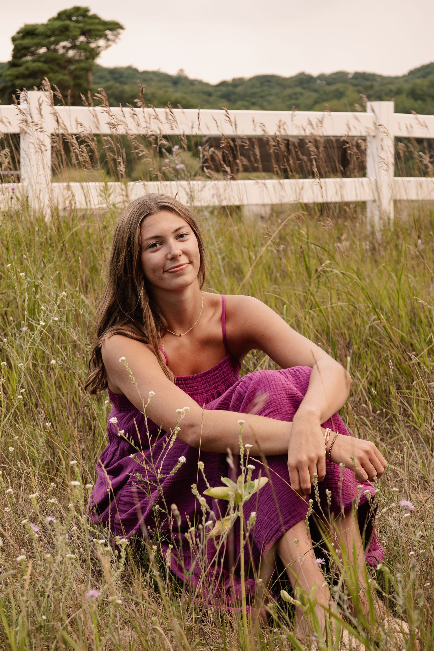 Young woman in purple outfit sitting in tall grass field near white wooden fence on overcast day.