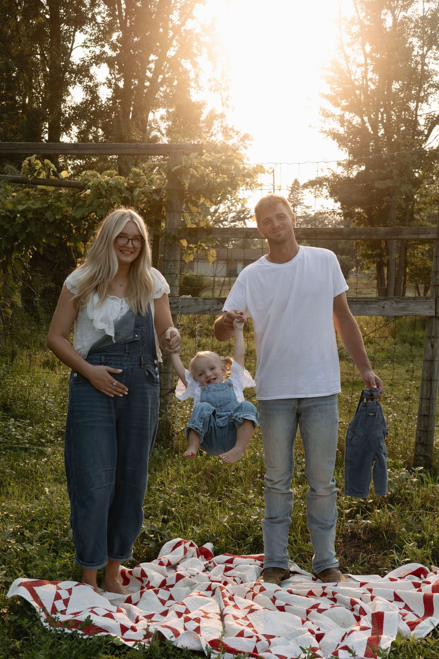 Young family with baby posing on a red quilt blanket outdoors at golden sunset hour.