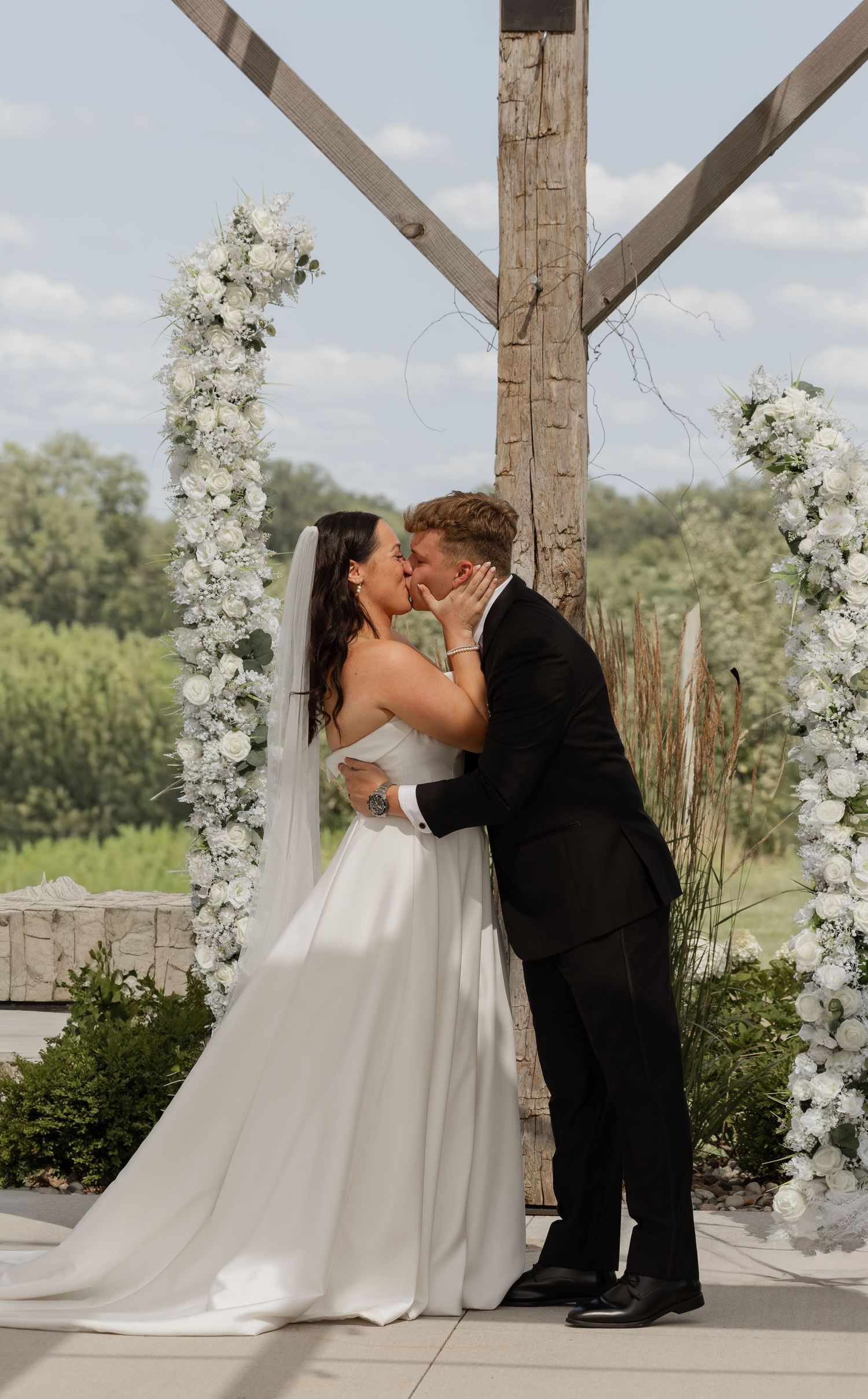 Bride and groom share first kiss at outdoor wedding altar adorned with white floral arrangements.