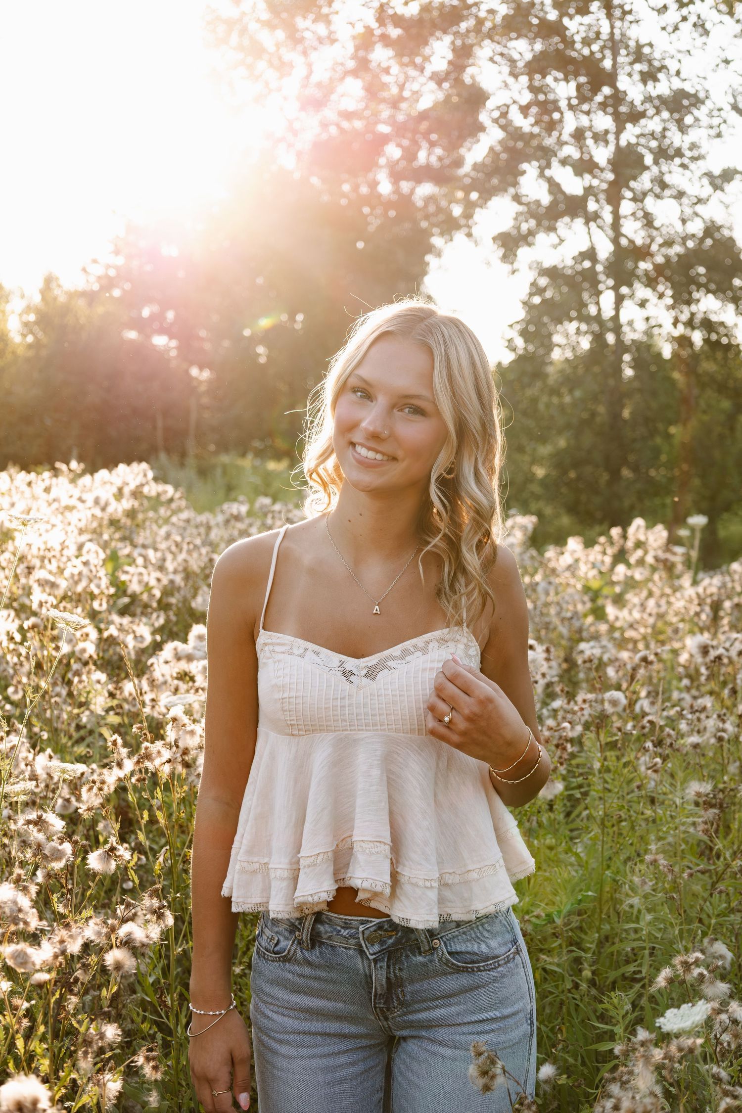 Smiling blonde woman in white lace crop top and jeans standing in wildflower field with golden sunset backlight.