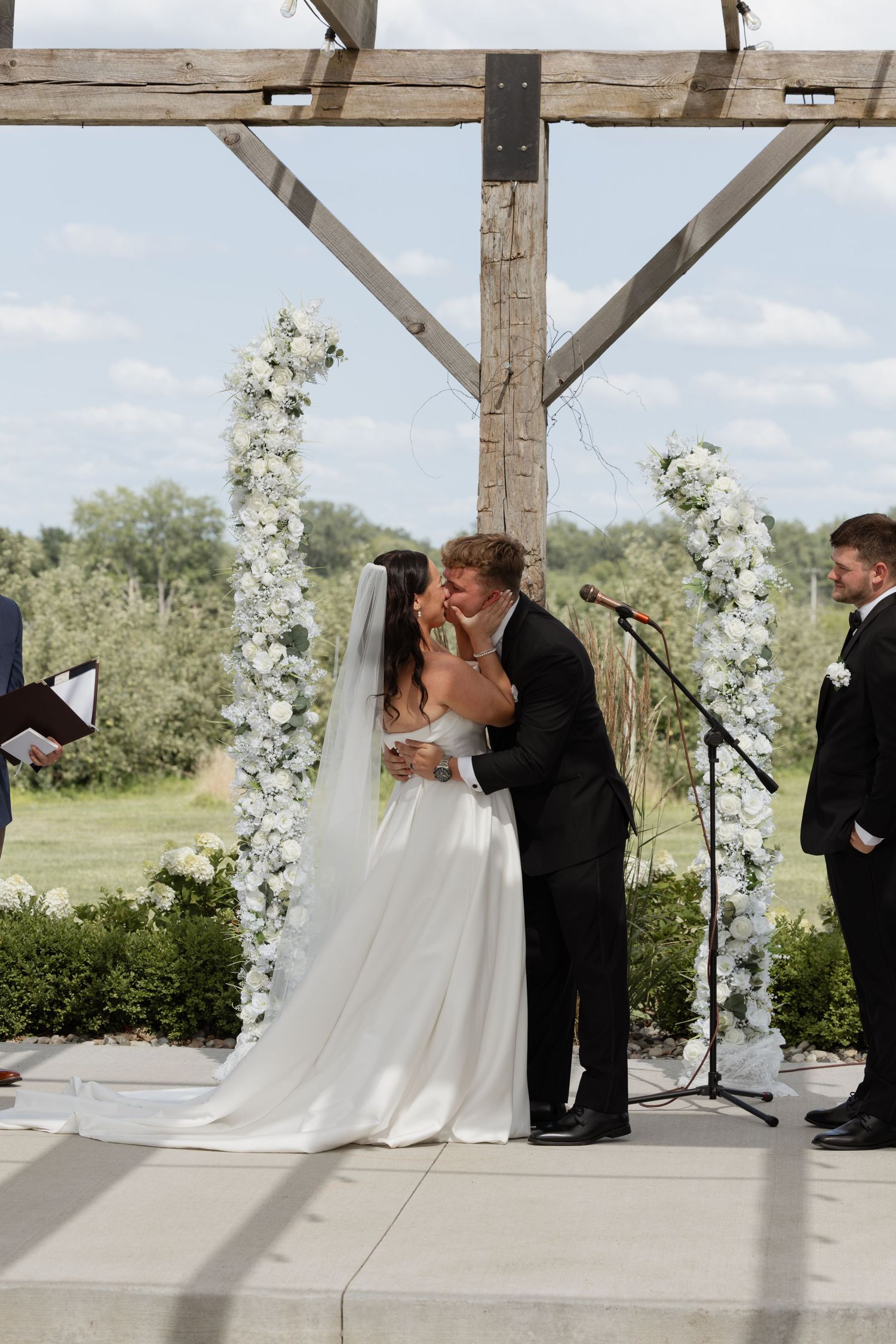 Bride and groom share first kiss at outdoor wedding ceremony under rustic wooden arch with white florals.