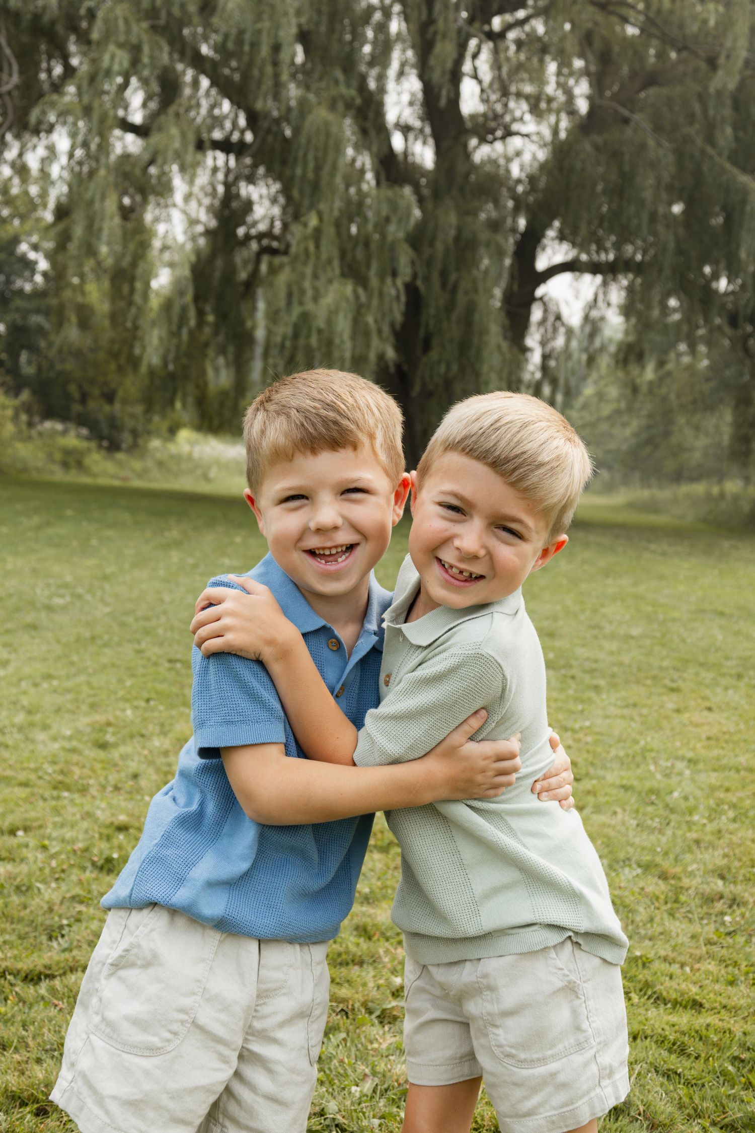 Two happy young boys hugging and smiling outdoors on green grass with a large willow tree in the background.