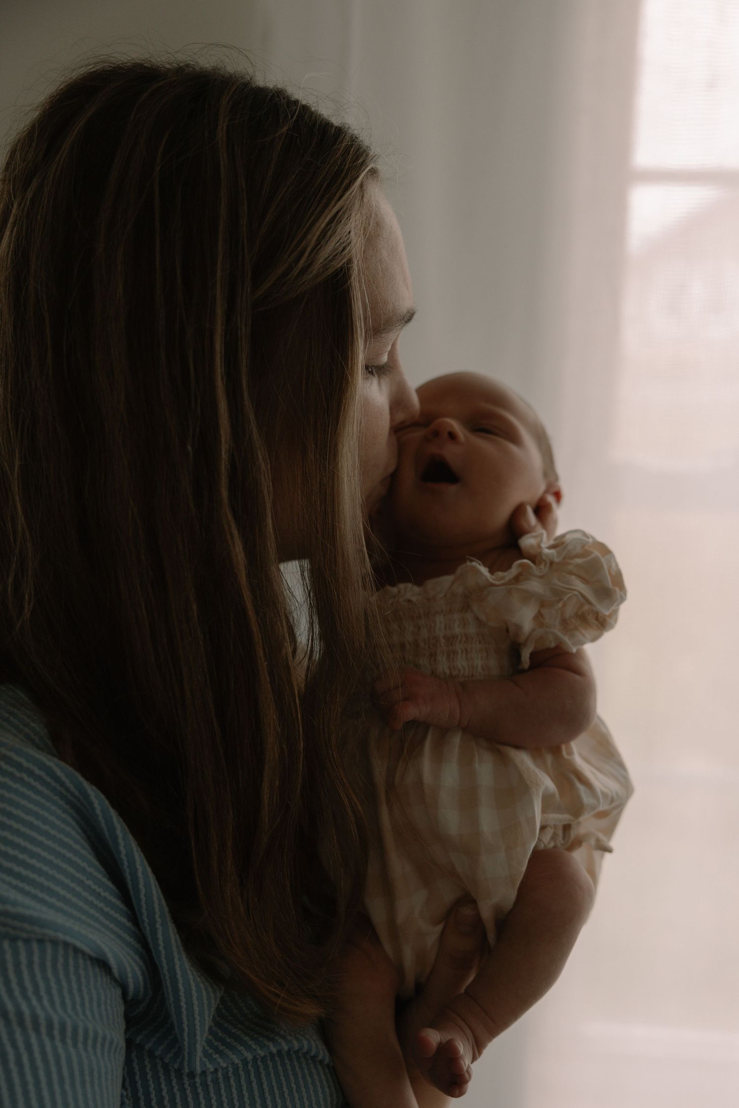 A mother tenderly holds her newborn baby near a window, sharing a gentle moment in soft, warm light.