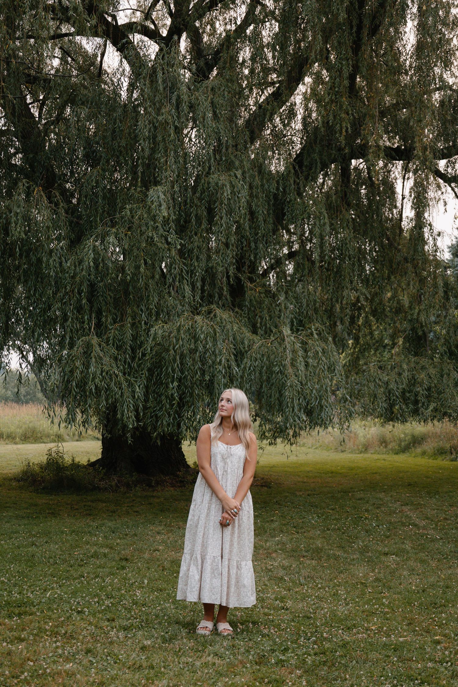 A young blonde woman in a white midi dress stands gracefully beneath a large weeping willow tree on a lush green lawn.