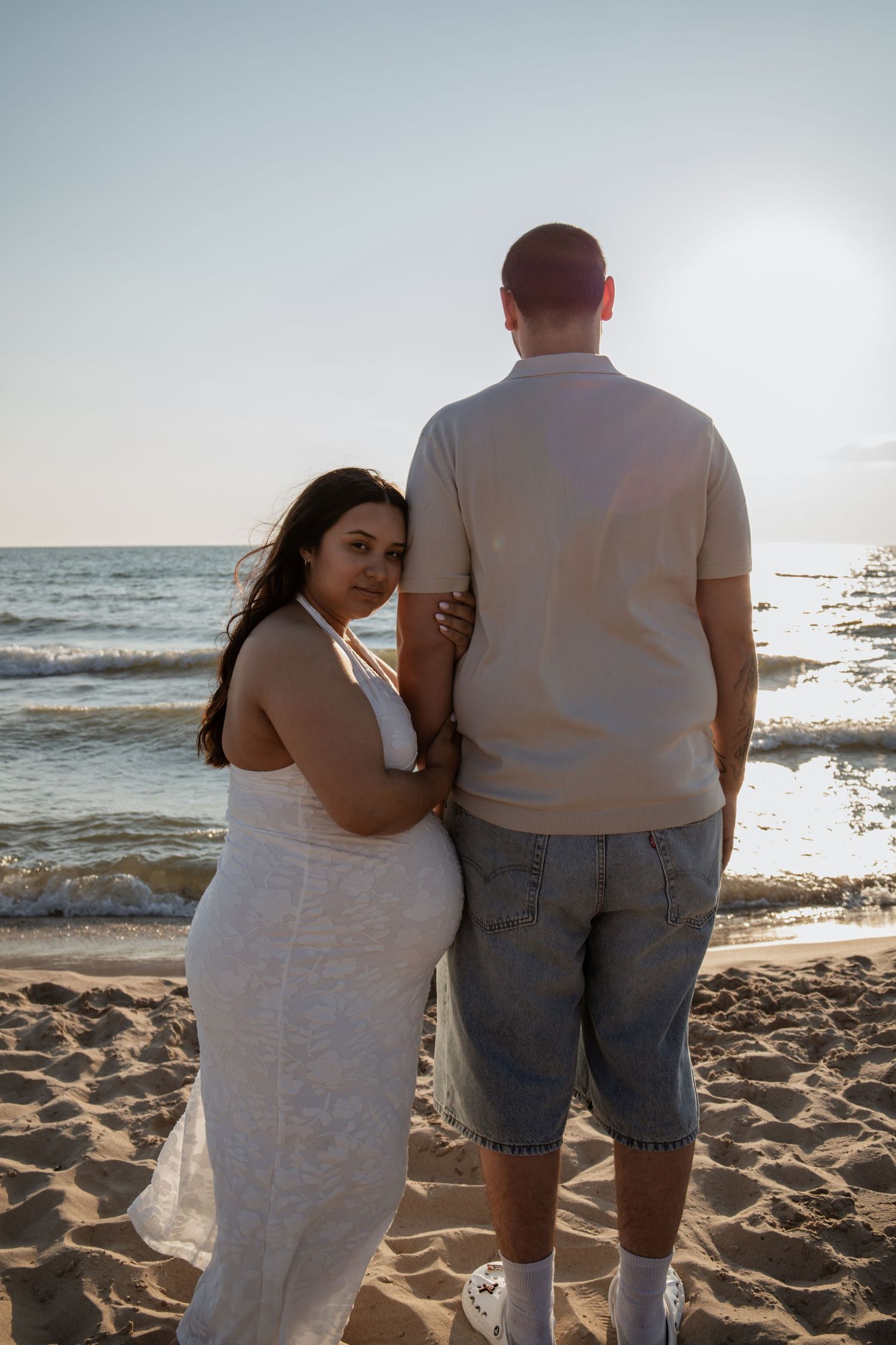 Couple standing together on a sandy beach at sunset, woman in white dress looking back at camera.