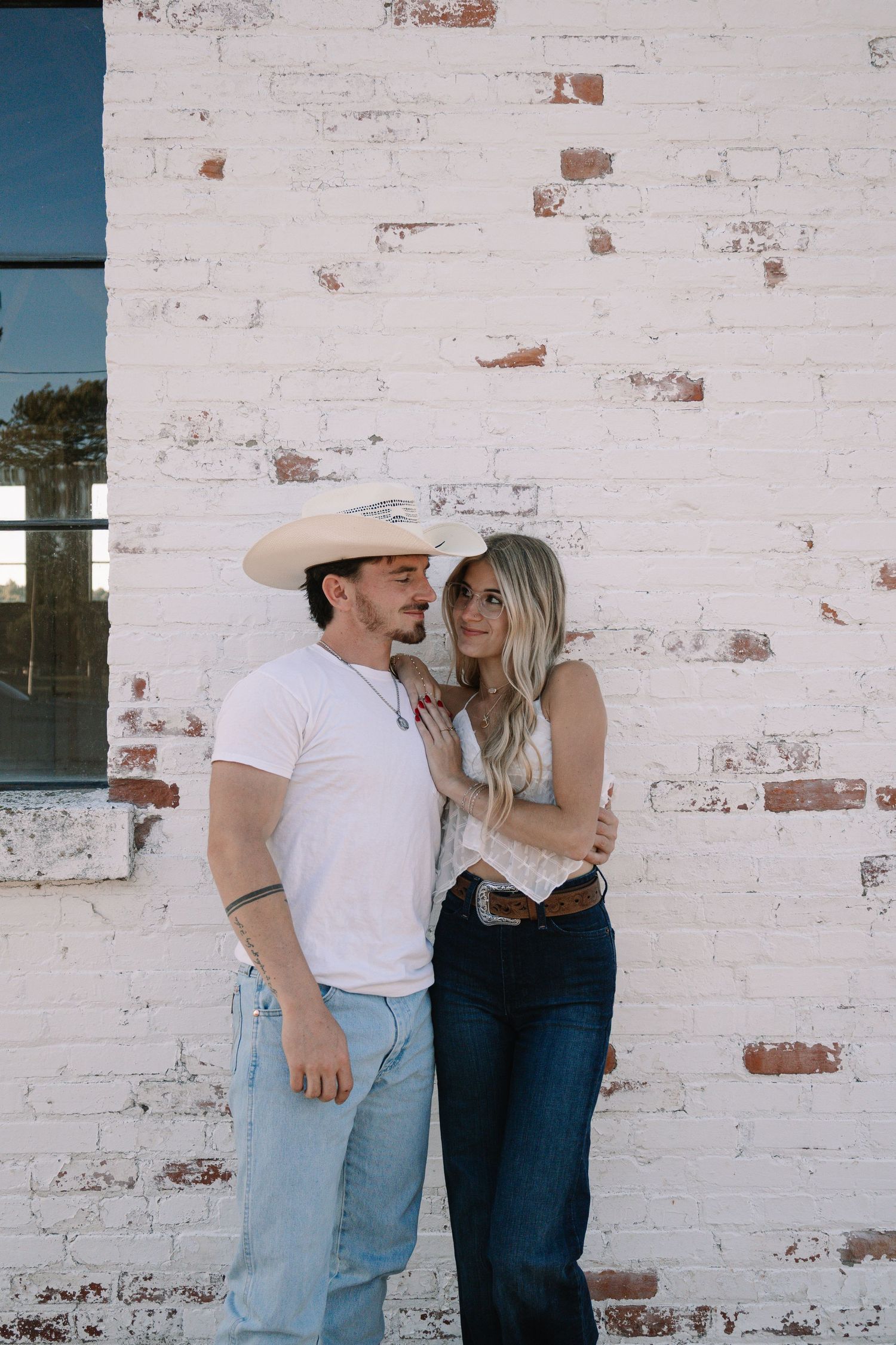 A couple poses romantically against a white brick wall, man in cowboy hat and white tee, woman in jeans.